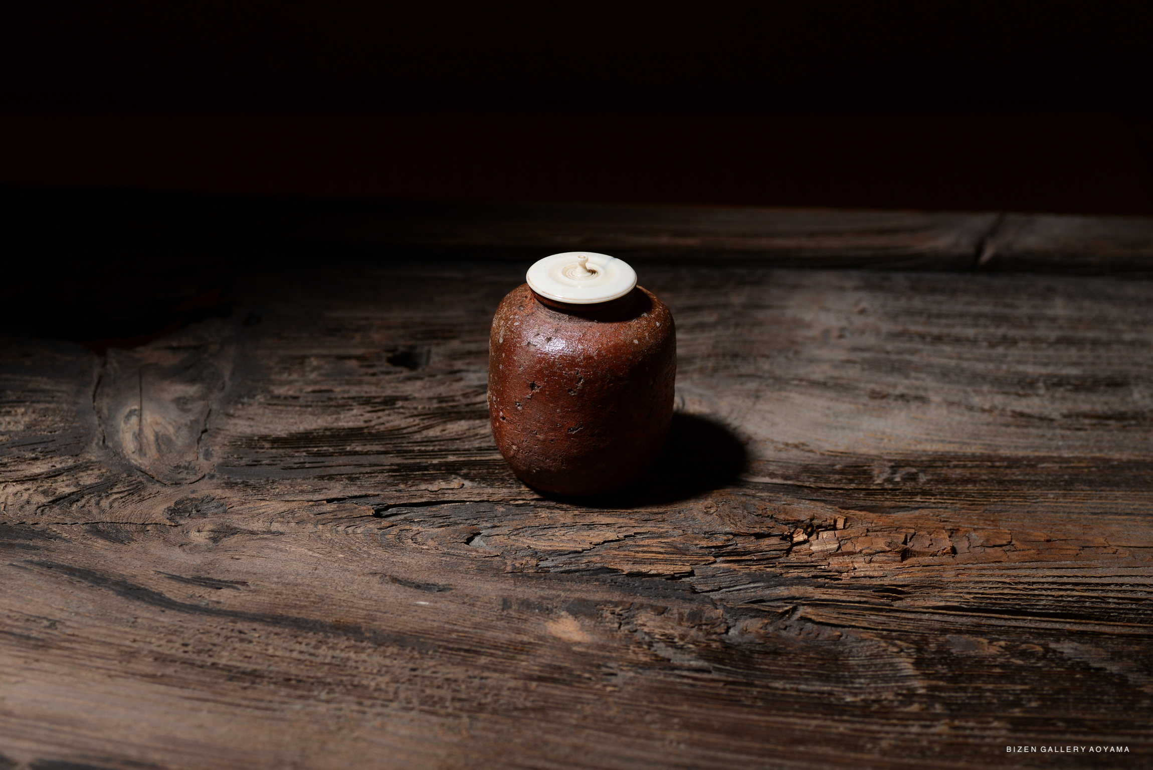 A Shigaraki Chaire with an engraved signature, featuring a round, reddish-brown pottery container resting on a wooden surface. The piece has a small white lid and exhibits signs of aging.