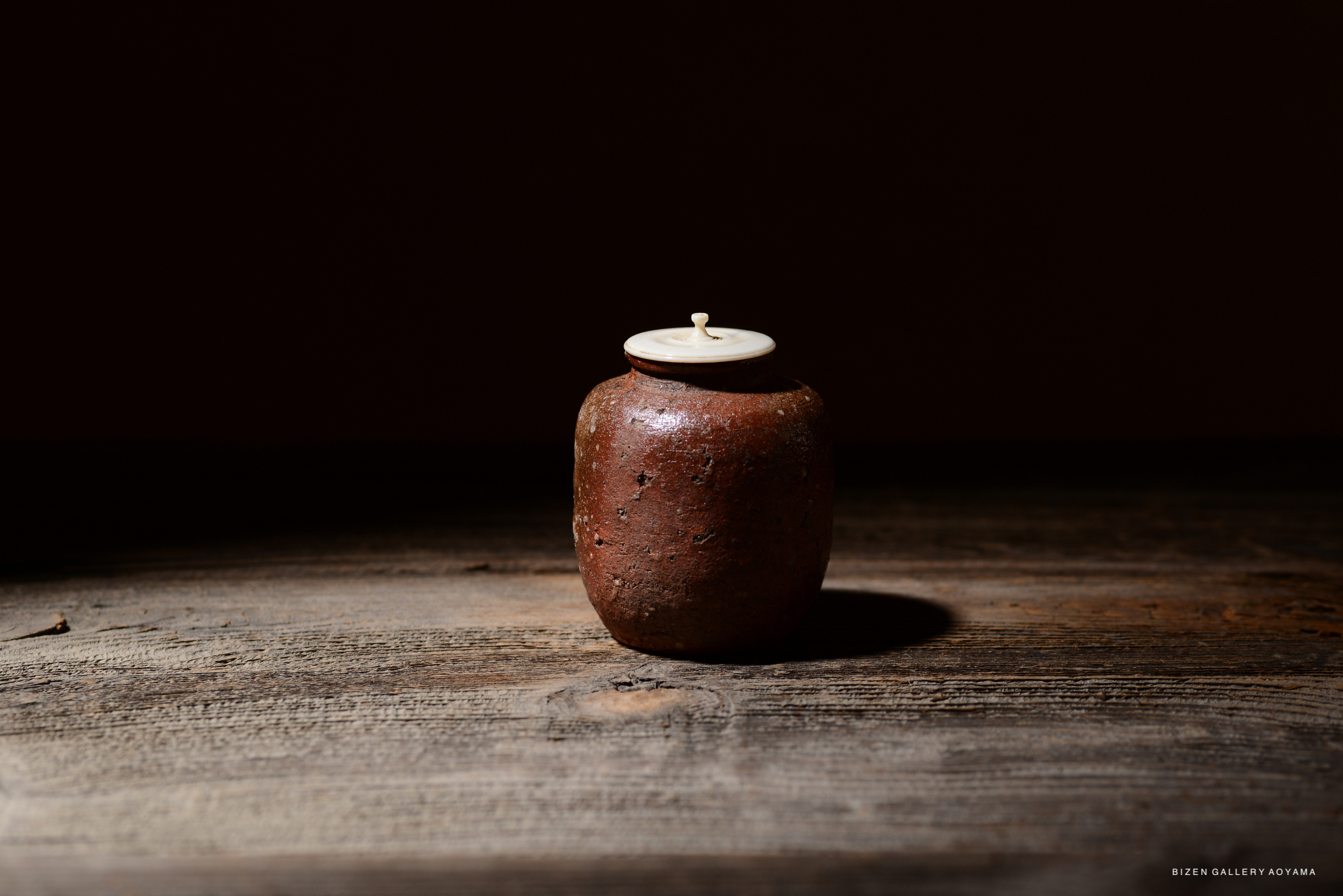 A rustic Shigaraki tea container with an engraved signature, featuring a simple round shape and a lightly colored lid, placed on a wooden surface with a dark background.