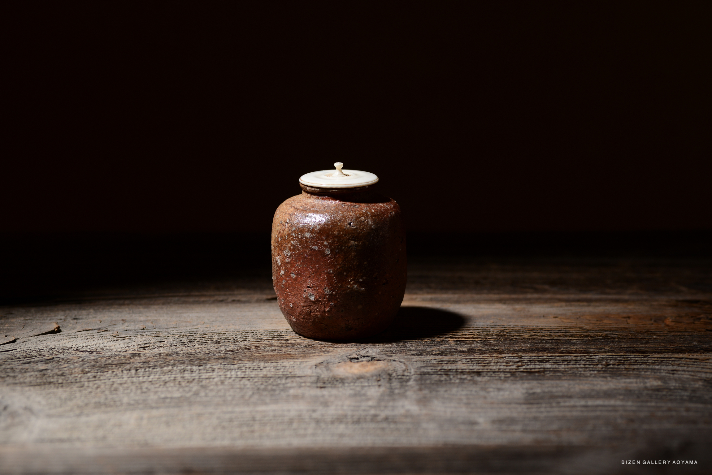 A rustic Shigaraki chaire tea container made of clay, featuring a light-colored lid, placed on a wooden surface with soft lighting.