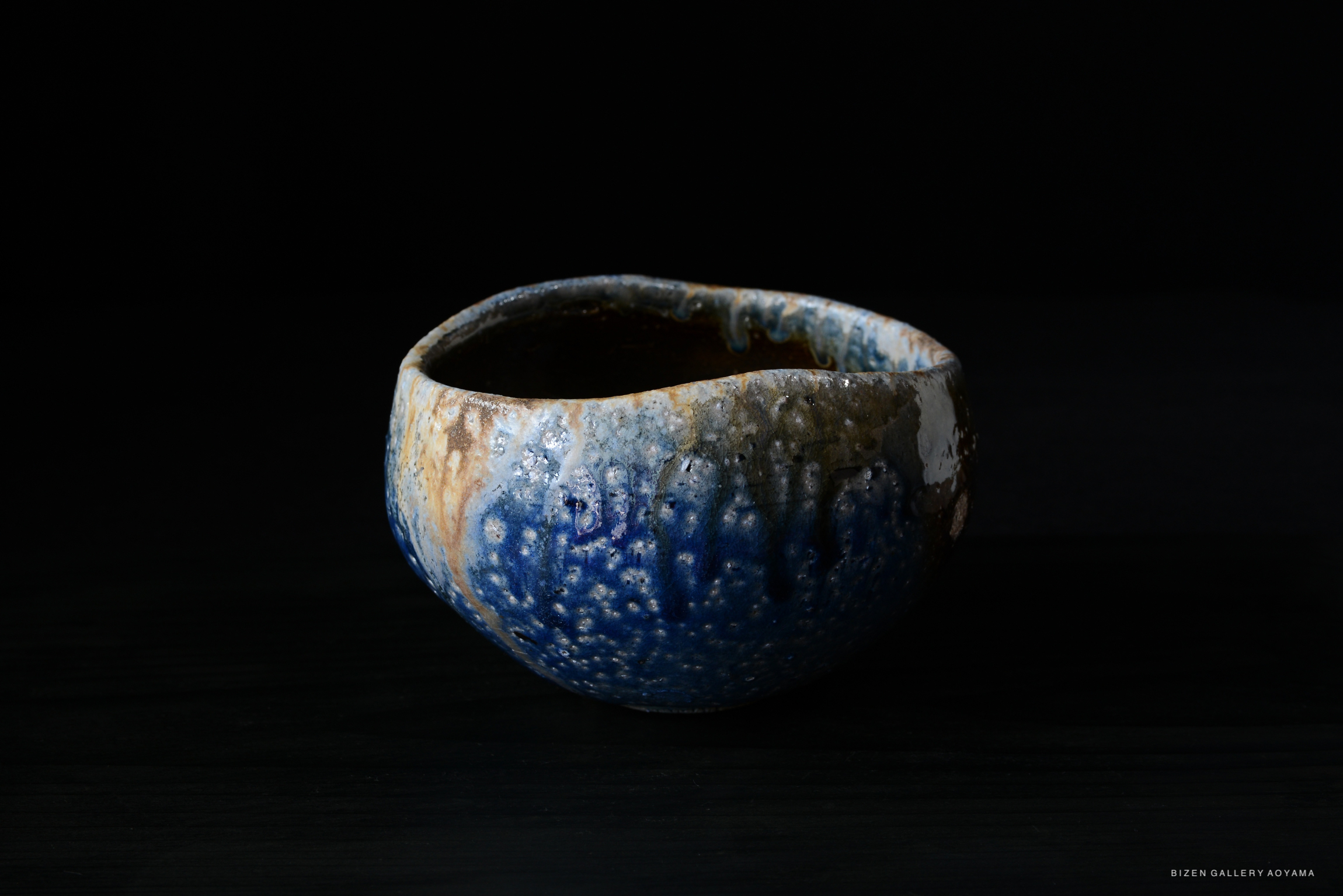 A uniquely shaped ceramic bowl with a textured surface, featuring a blend of blue and earthy tones, set against a black background.
