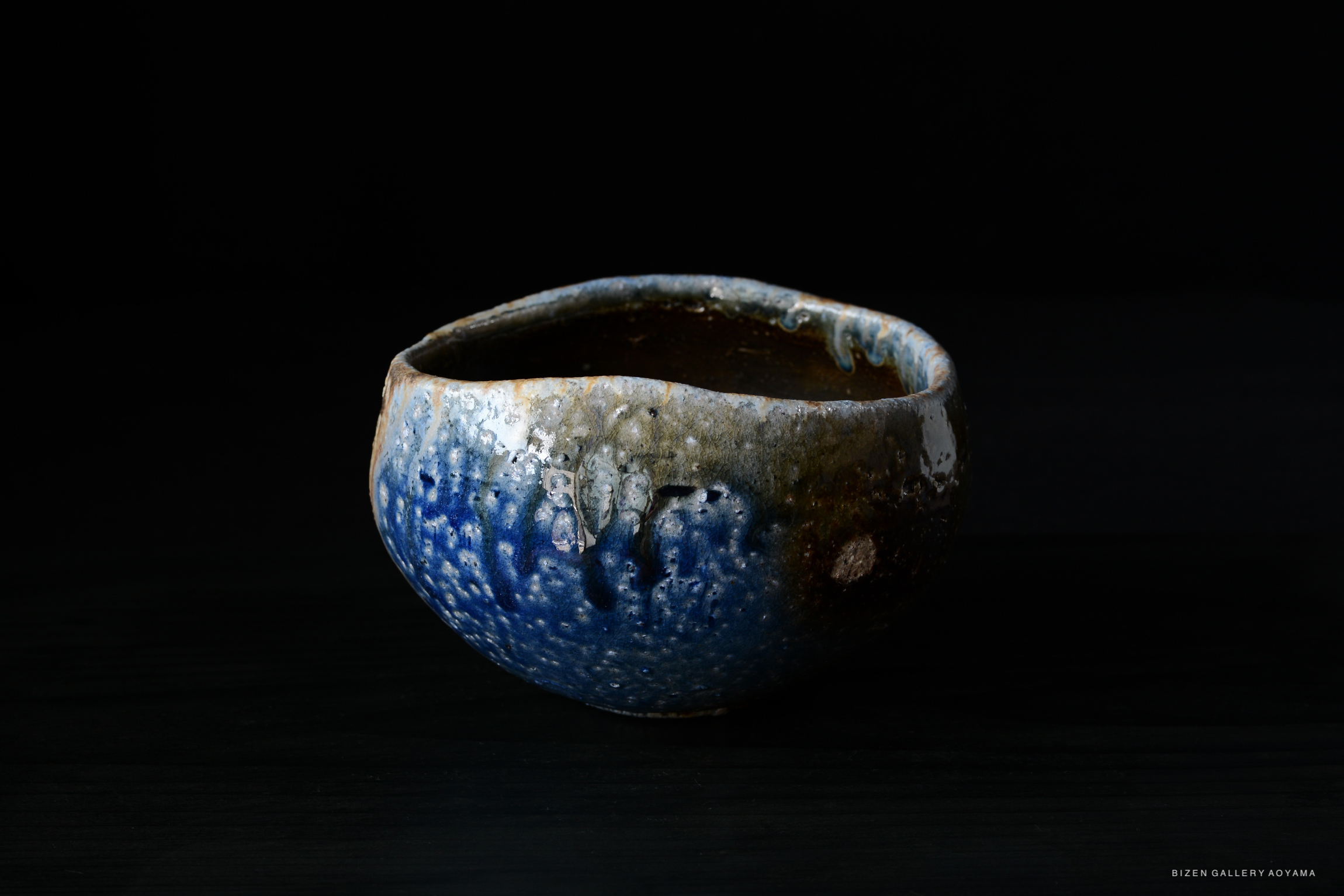A close-up view of a textured ceramic bowl with a blue and brown glaze against a black background.