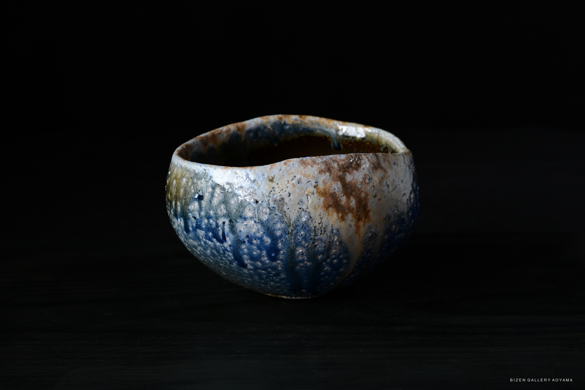 A handcrafted ceramic bowl with a textured surface and a blend of blue and brown glazes, set against a dark background.