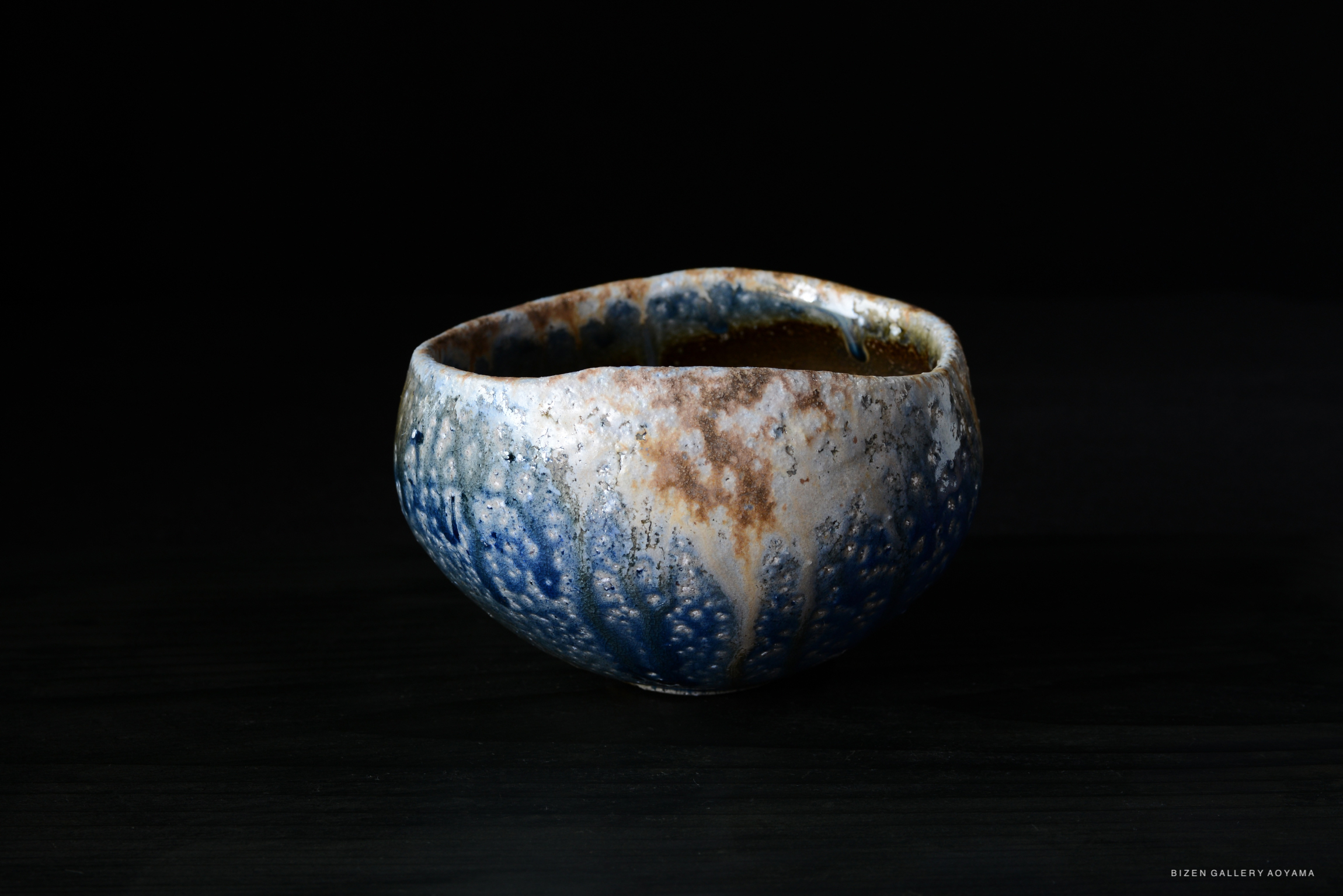 A handmade ceramic bowl with a textured surface featuring blue and brown glazes, placed on a dark table.