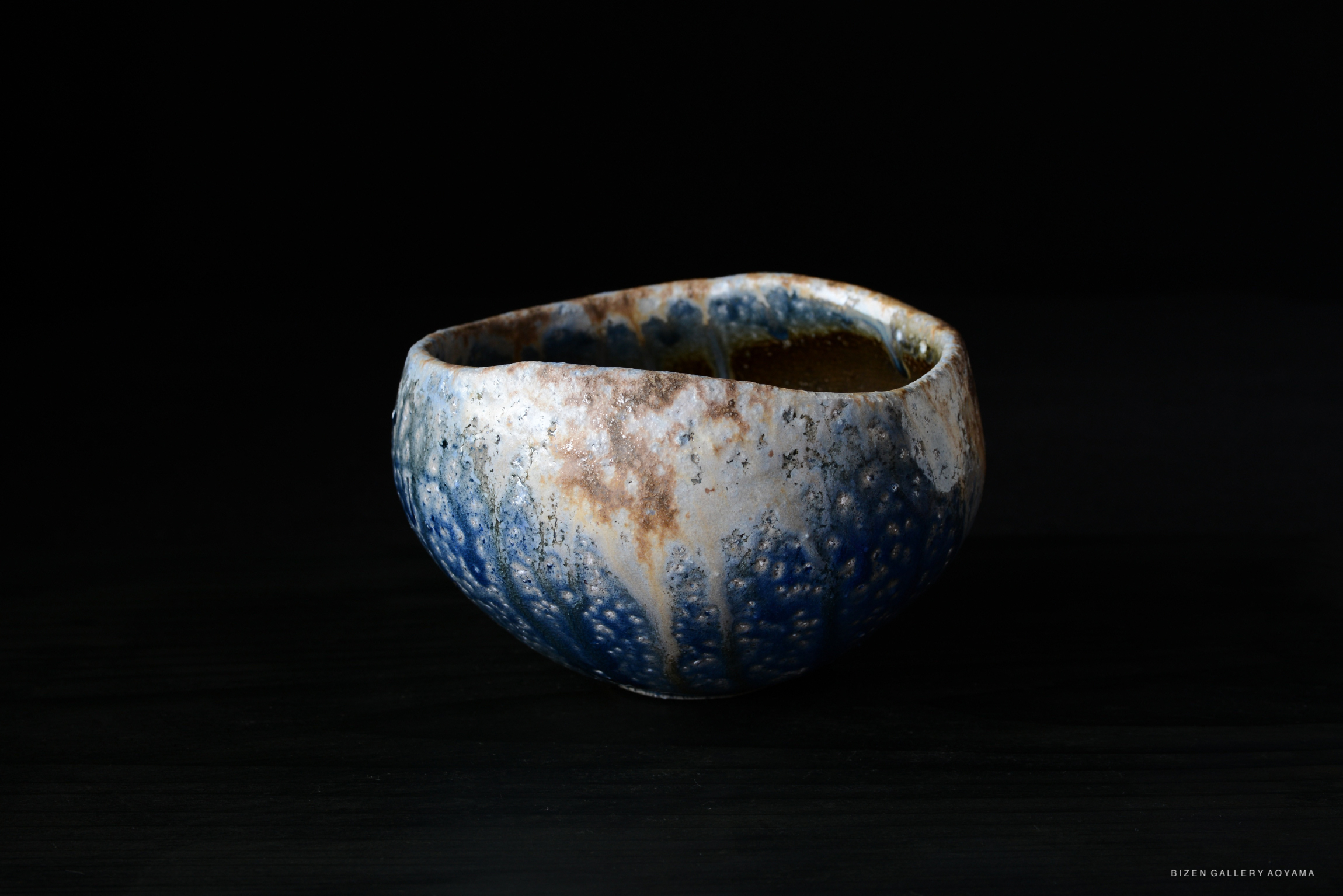 A unique, handcrafted ceramic bowl featuring a textured surface with blue and earthy tones, set against a dark background.