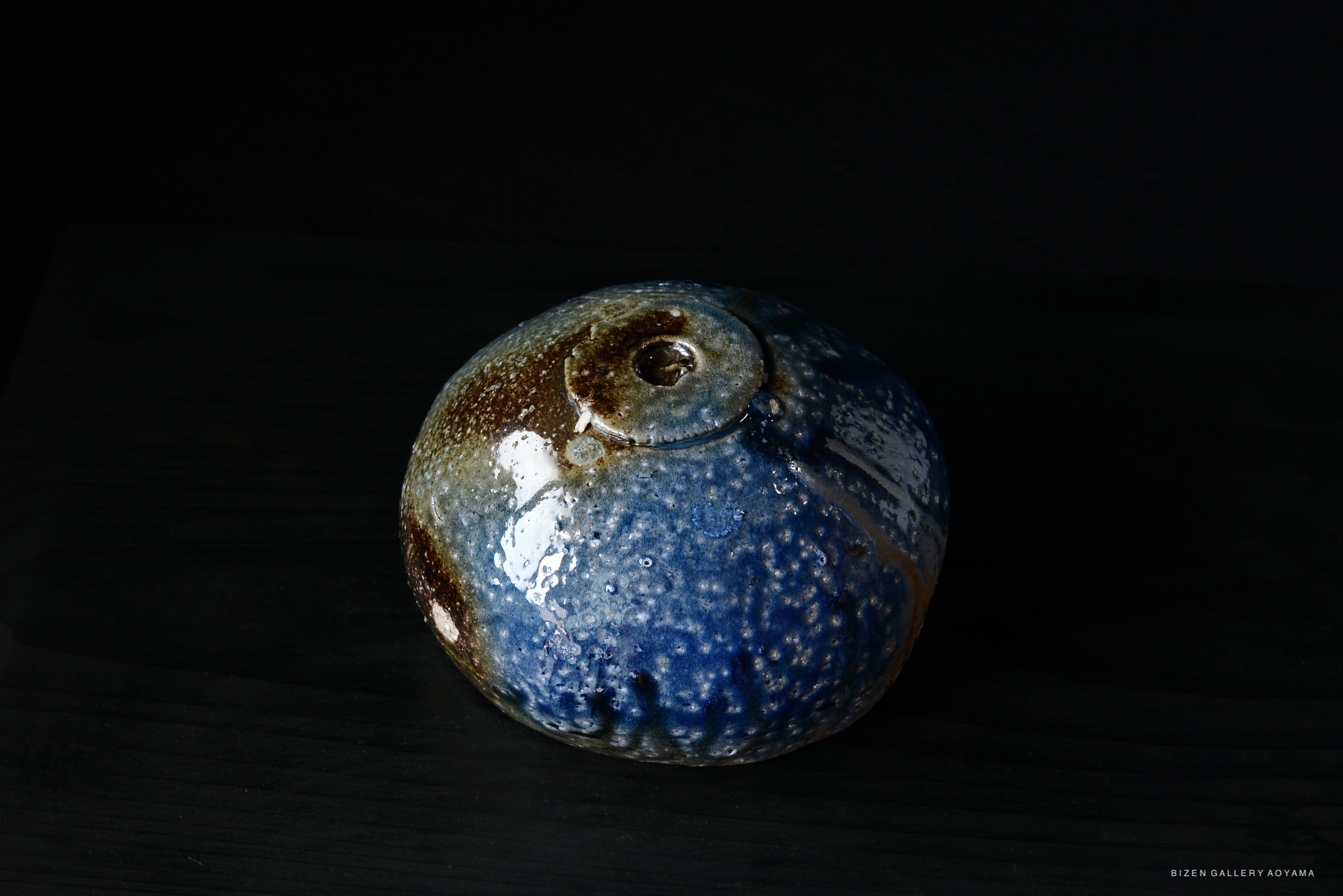Close-up of a round ceramic vase with a glossy blue and brown glaze, placed on a dark surface.