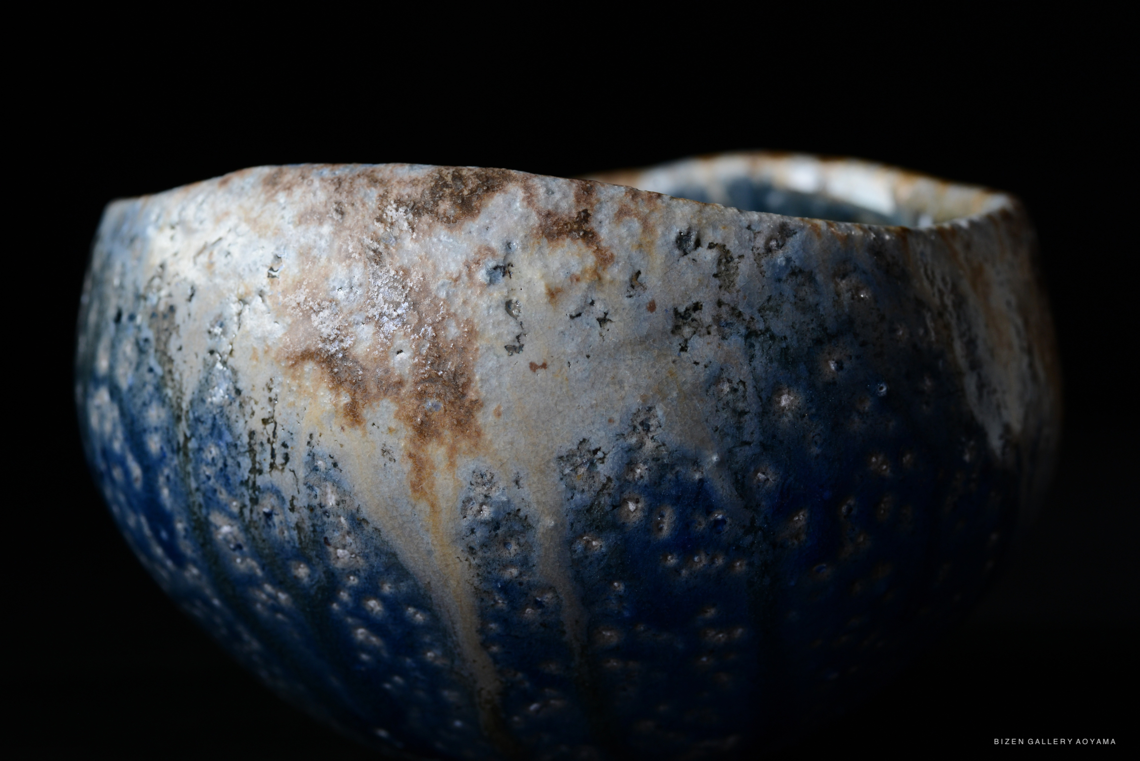 Close-up of a textured ceramic bowl with a blue and earthy color palette, reflecting light against a dark background.