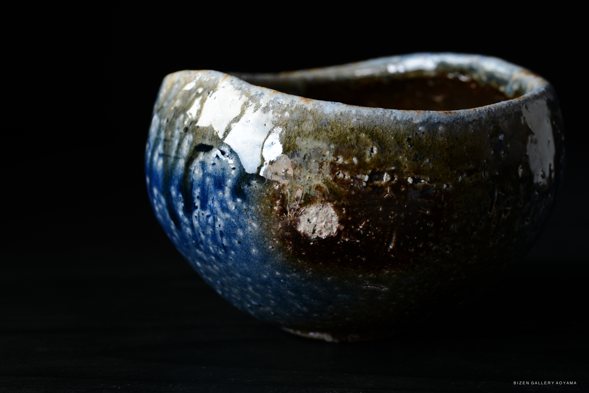 A close-up of a handcrafted ceramic bowl featuring a glossy blue and brown glaze, with textured details and an organic shape, set against a black background.