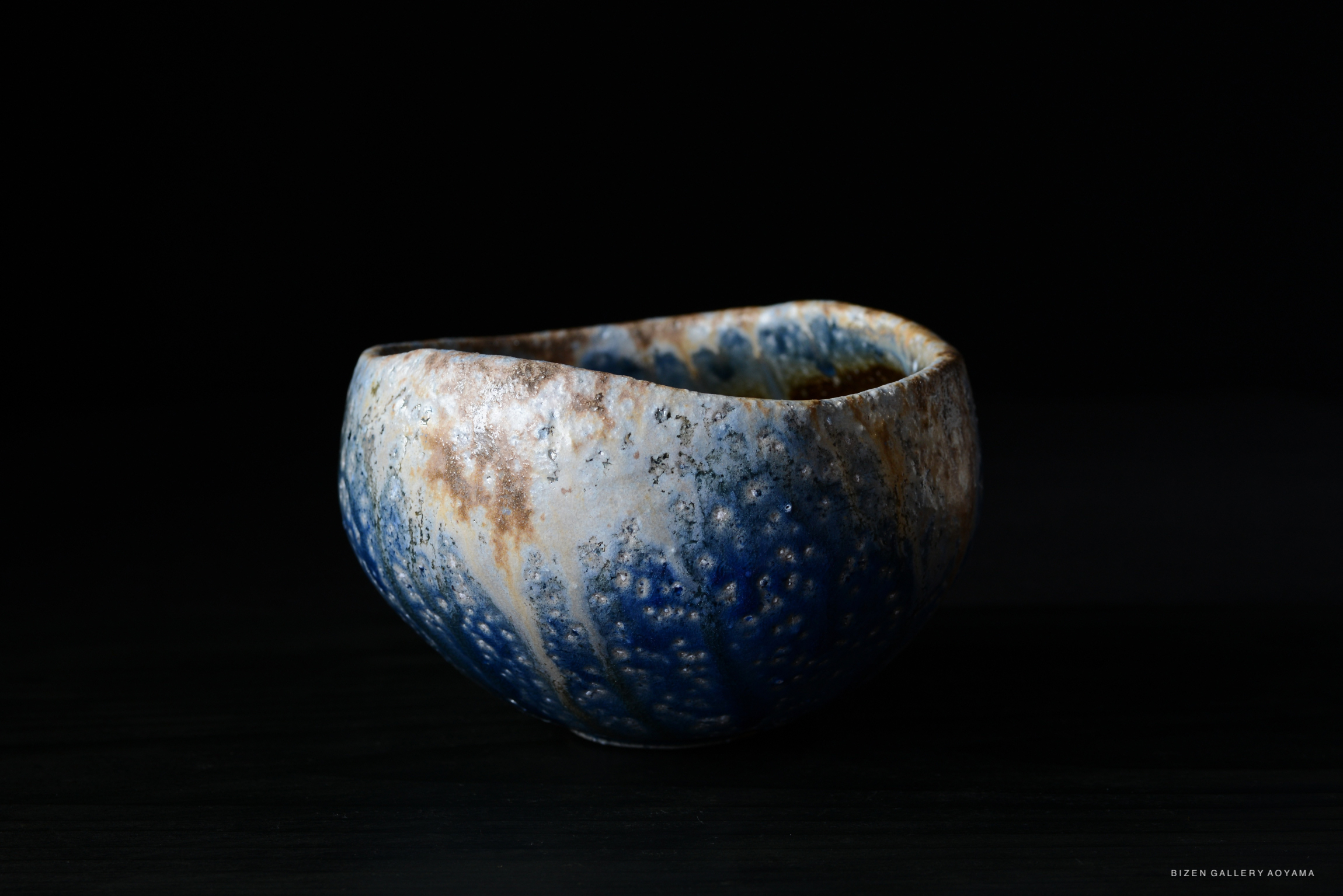 A handcrafted ceramic bowl with a textured surface featuring blue and brown hues, shown against a dark background.