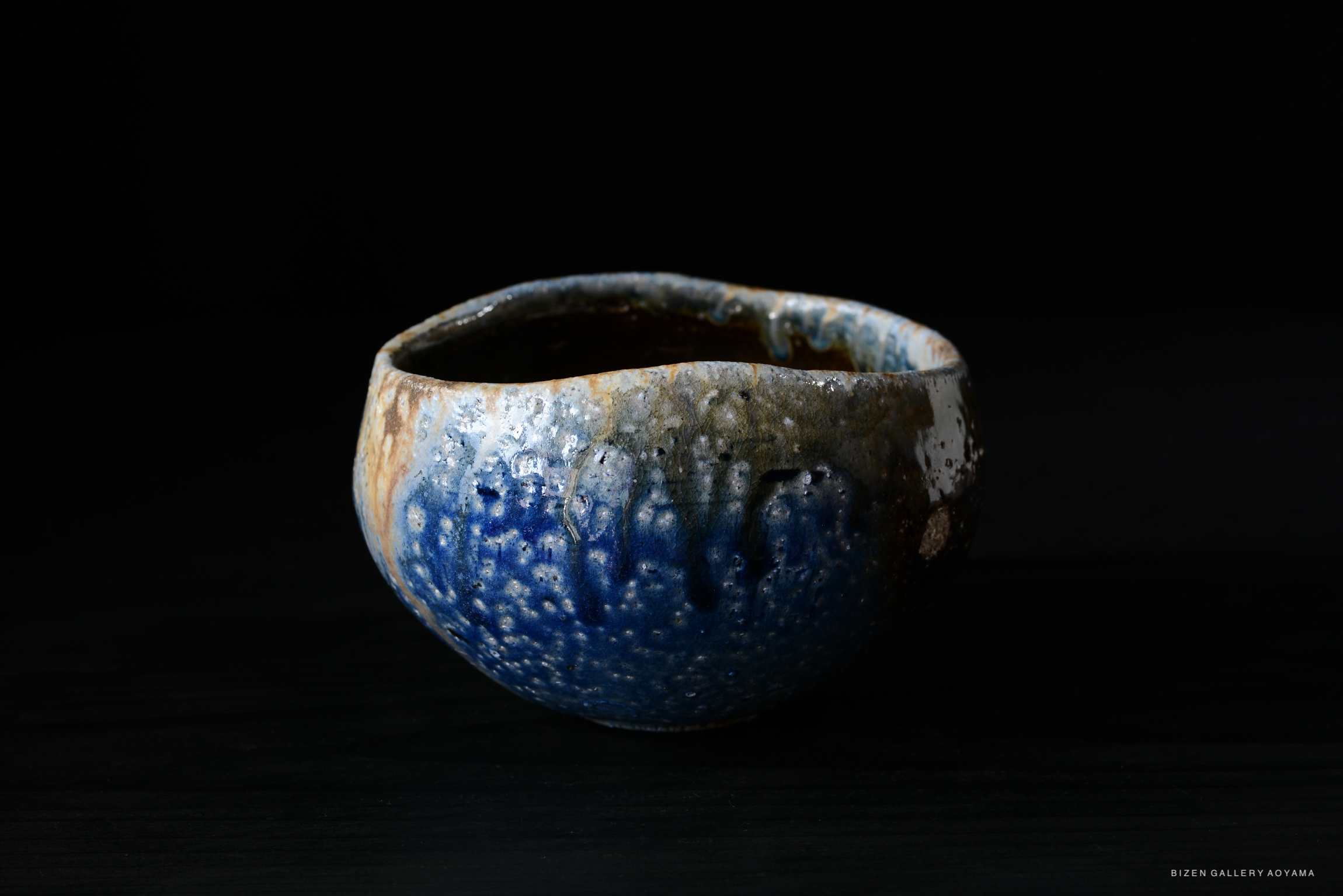 A handcrafted ceramic bowl with a mix of blue and earthy tones, featuring a textured surface and a rustic appearance, set against a dark background.