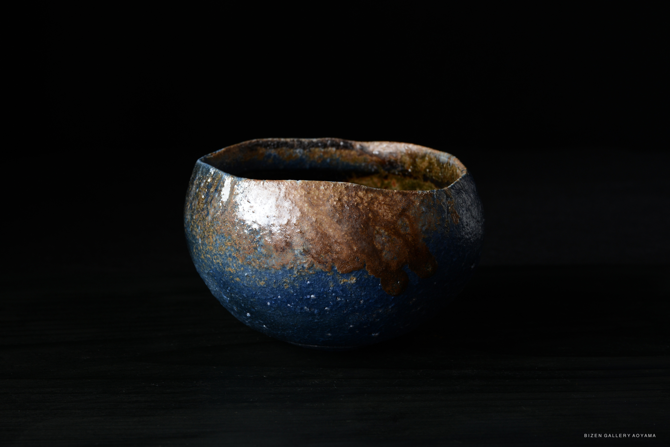 A close-up view of a handmade pottery bowl with a glossy blue and brown finish, showcasing unique textures and colors against a dark background.