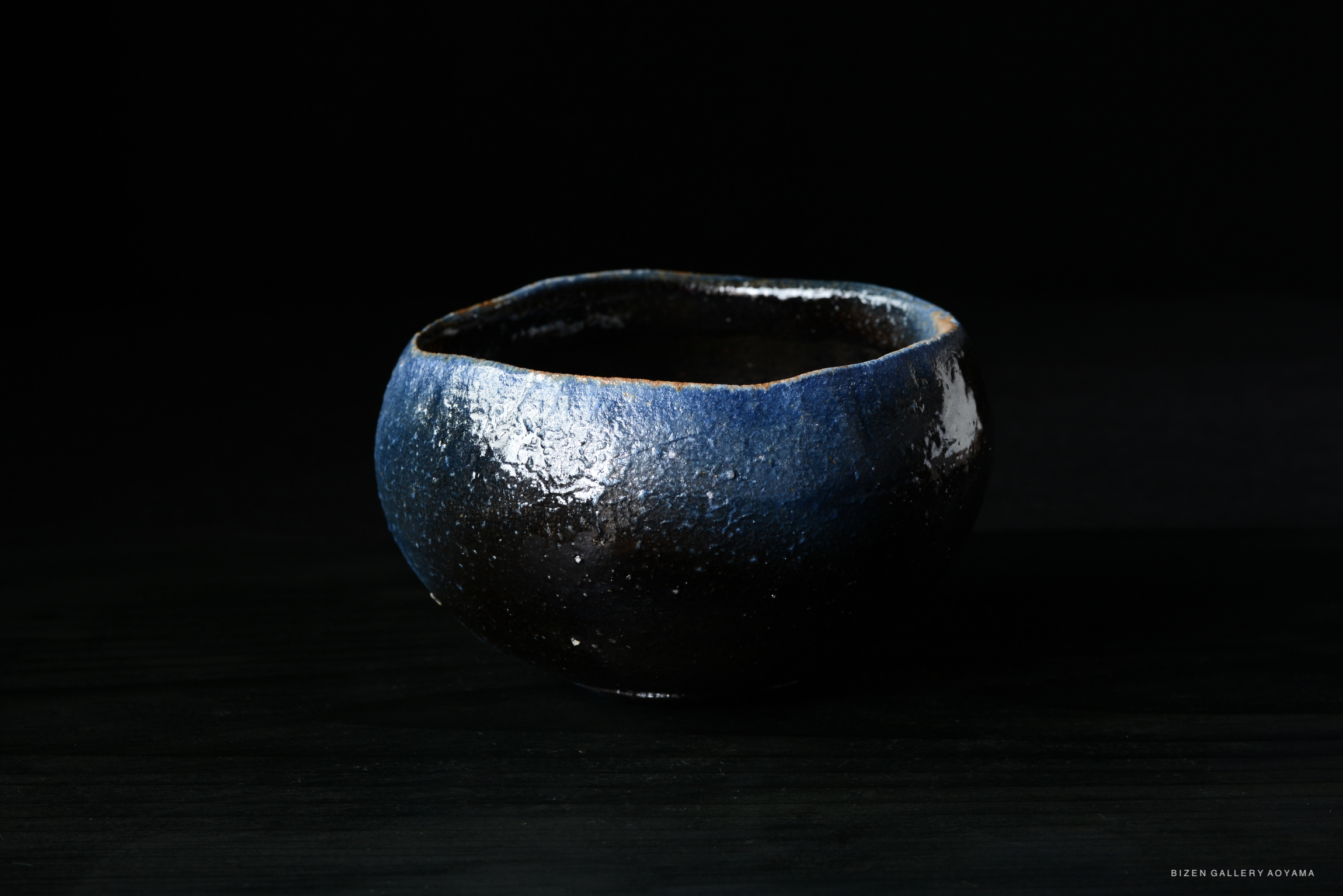 A handcrafted ceramic bowl featuring a textured black surface with a blue rim, set against a dark background.