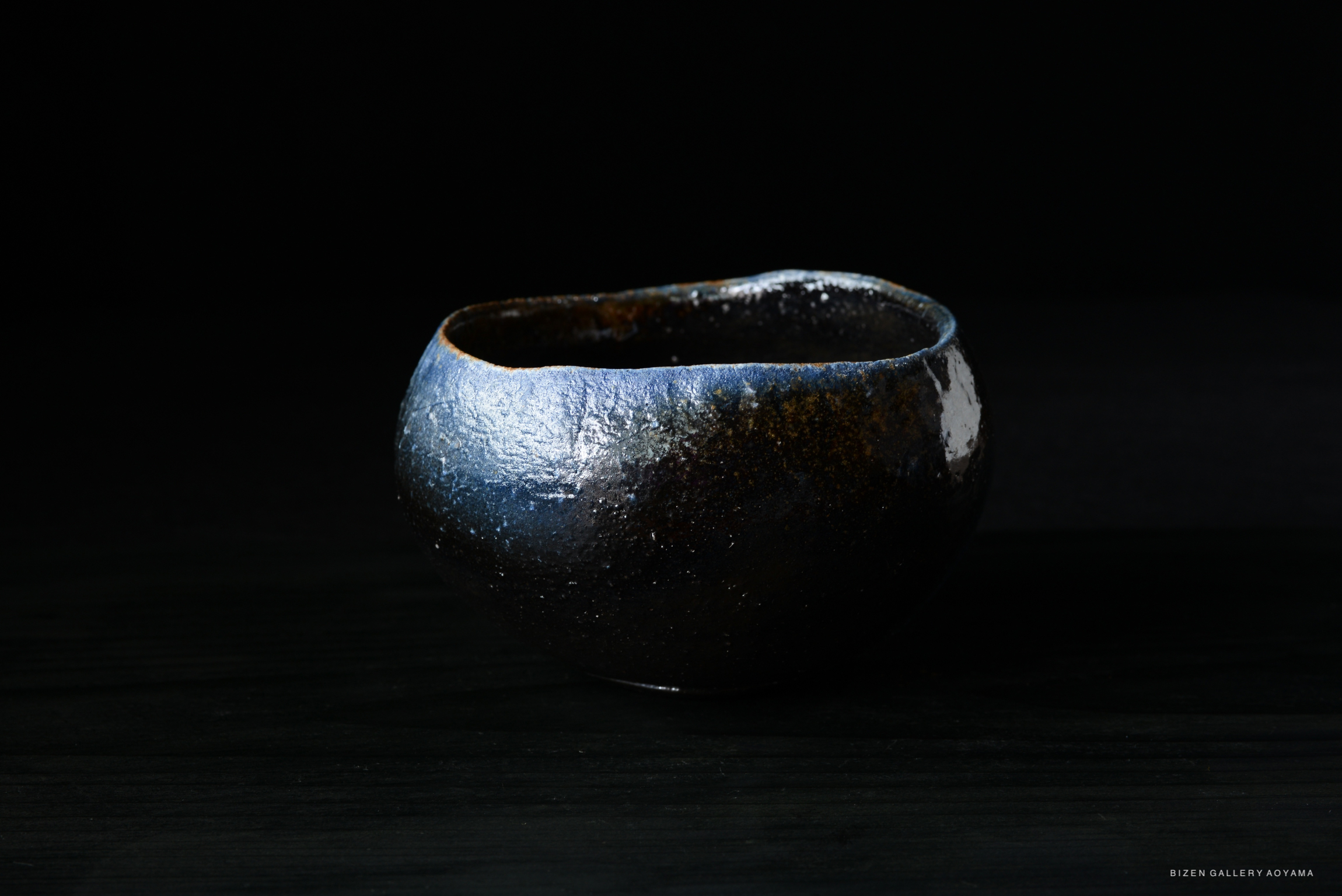 A round, textured ceramic bowl with a dark blue and metallic finish, set against a black background.