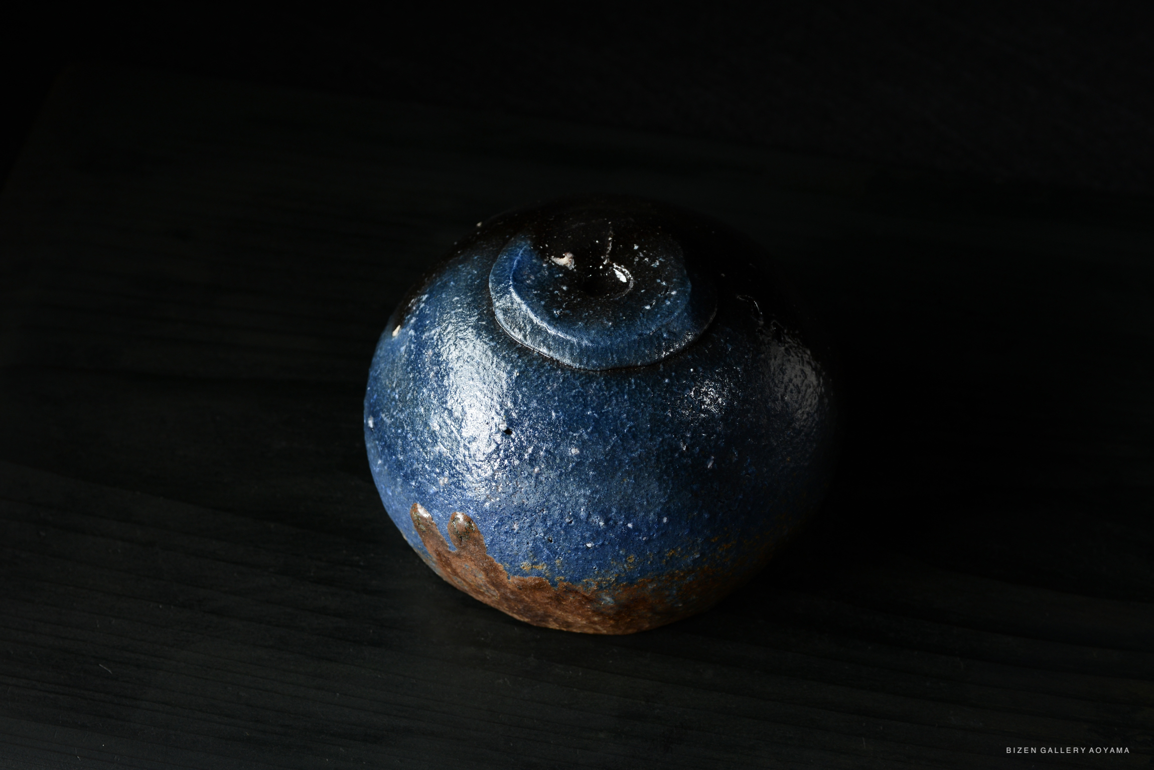 A round, blue ceramic piece with a glossy finish, featuring a smooth top and a rustic brown base, set against a dark background.