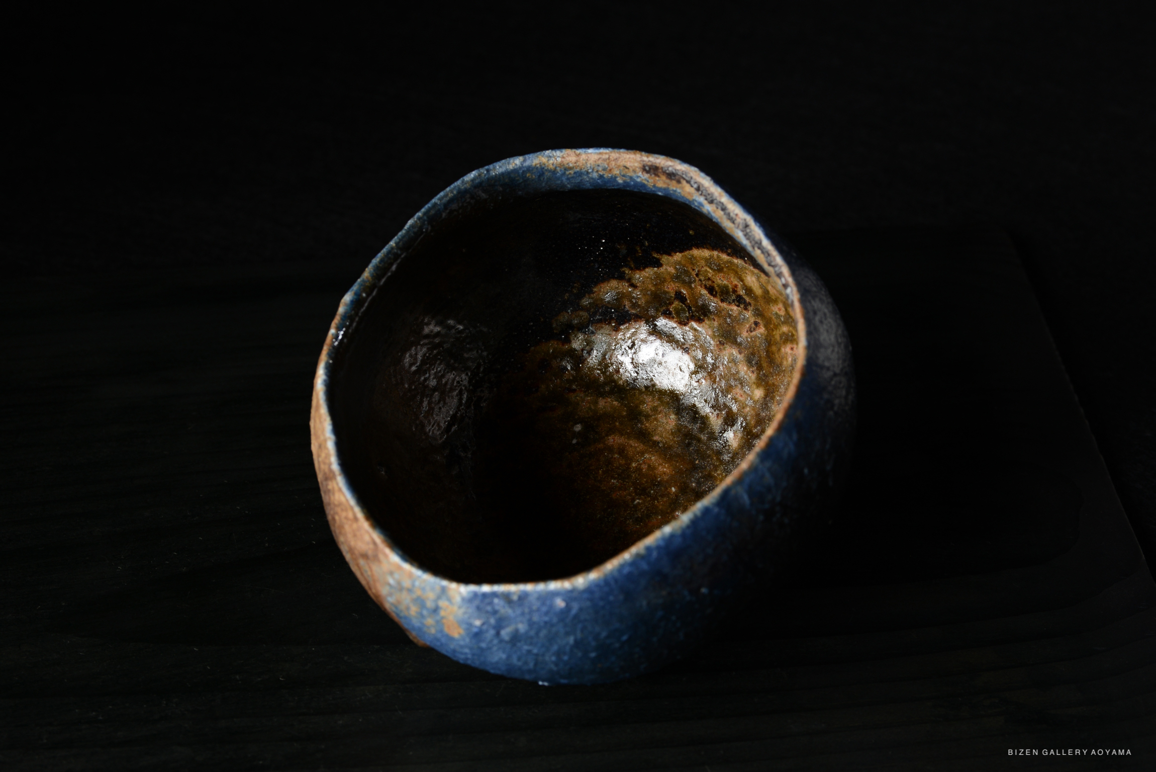 A top-down view of a handcrafted ceramic bowl with a blue rim and a textured dark interior, set against a dark background.
