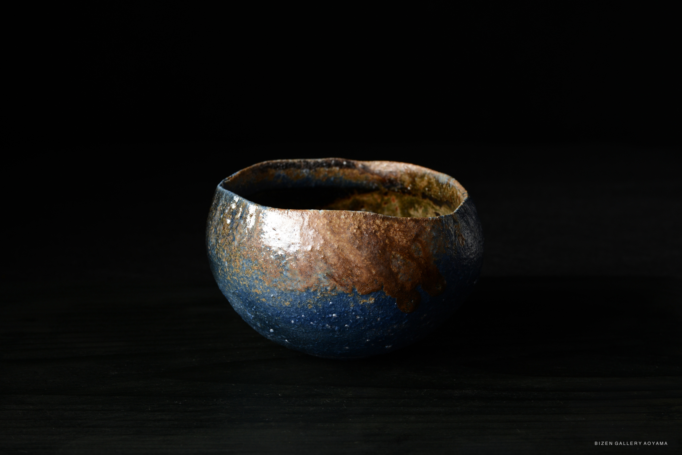 A close-up of a unique blue and brown glazed ceramic bowl with a textured surface, set against a dark background.