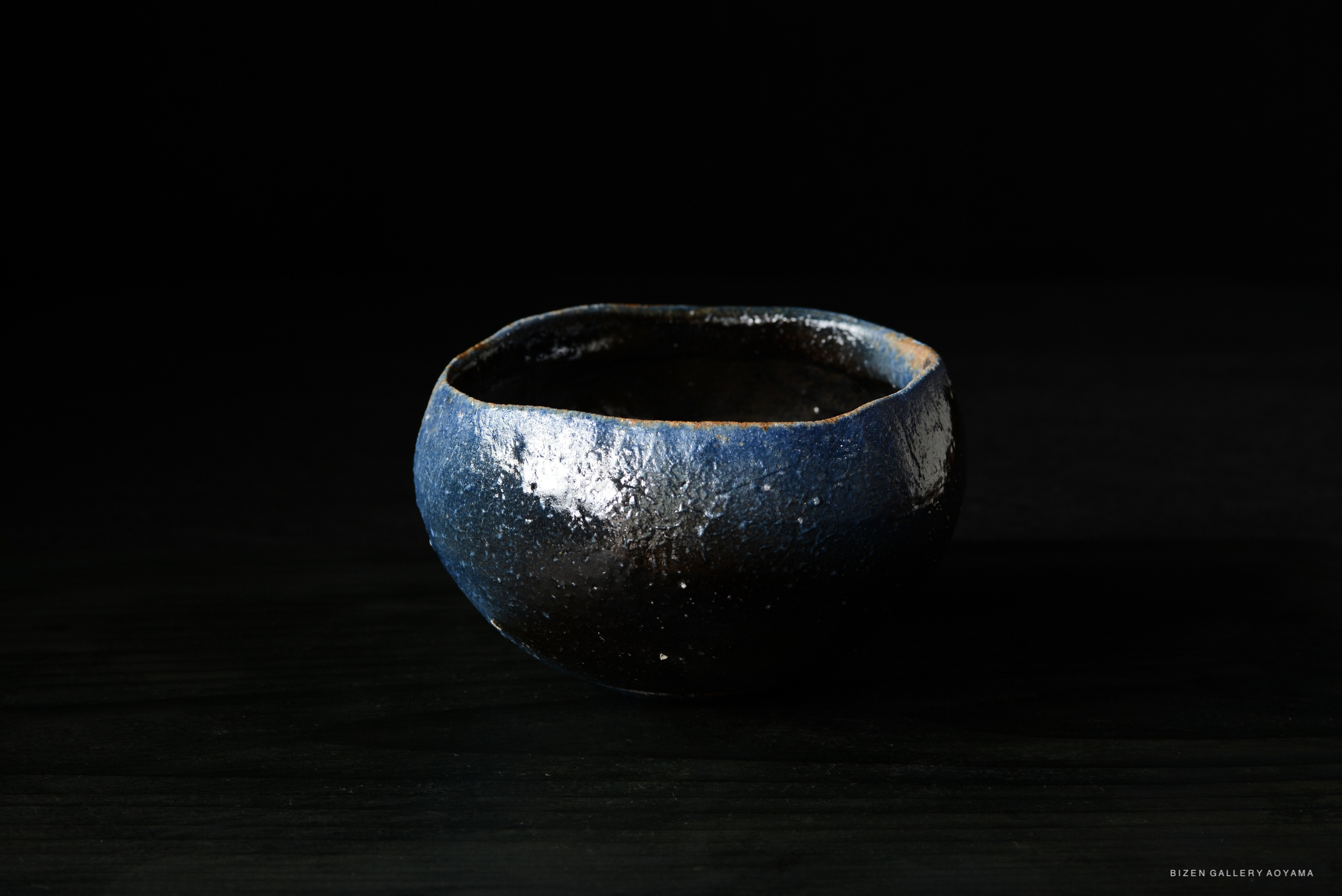 A close-up of a small, handcrafted ceramic bowl with a glossy blue-black finish, set against a dark background.