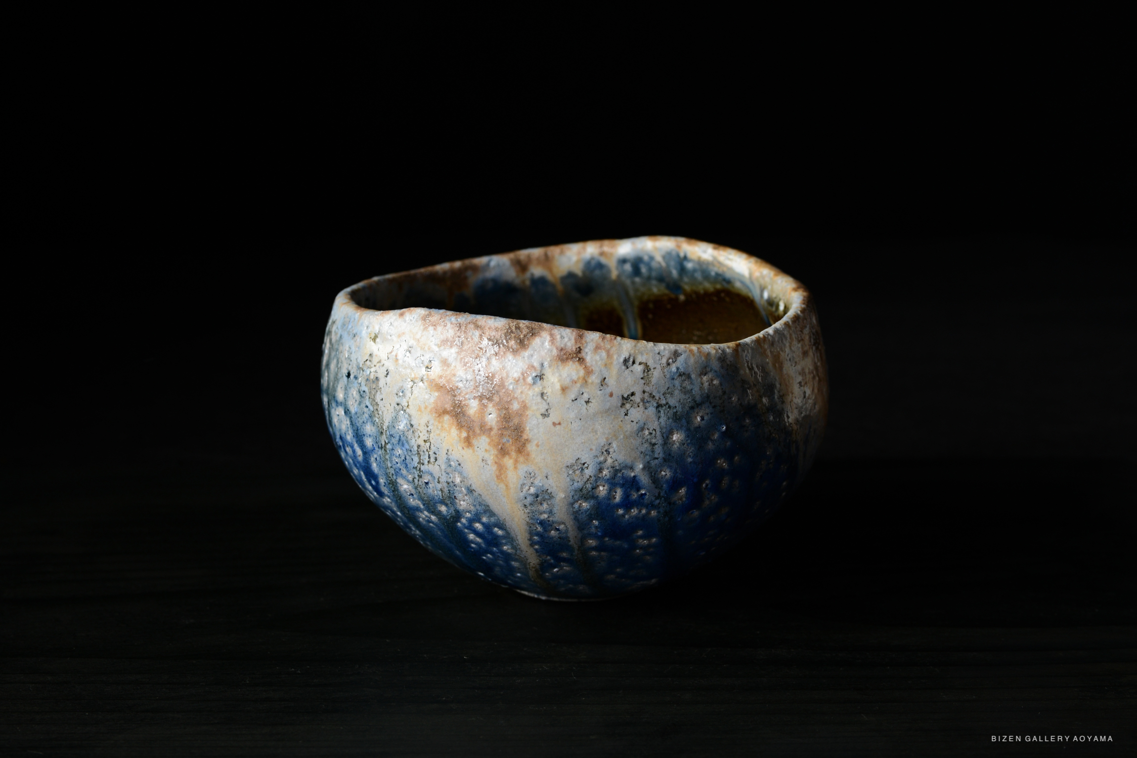 Close-up of a uniquely shaped ceramic bowl with a textured surface and a blue gradient coloration against a dark background.