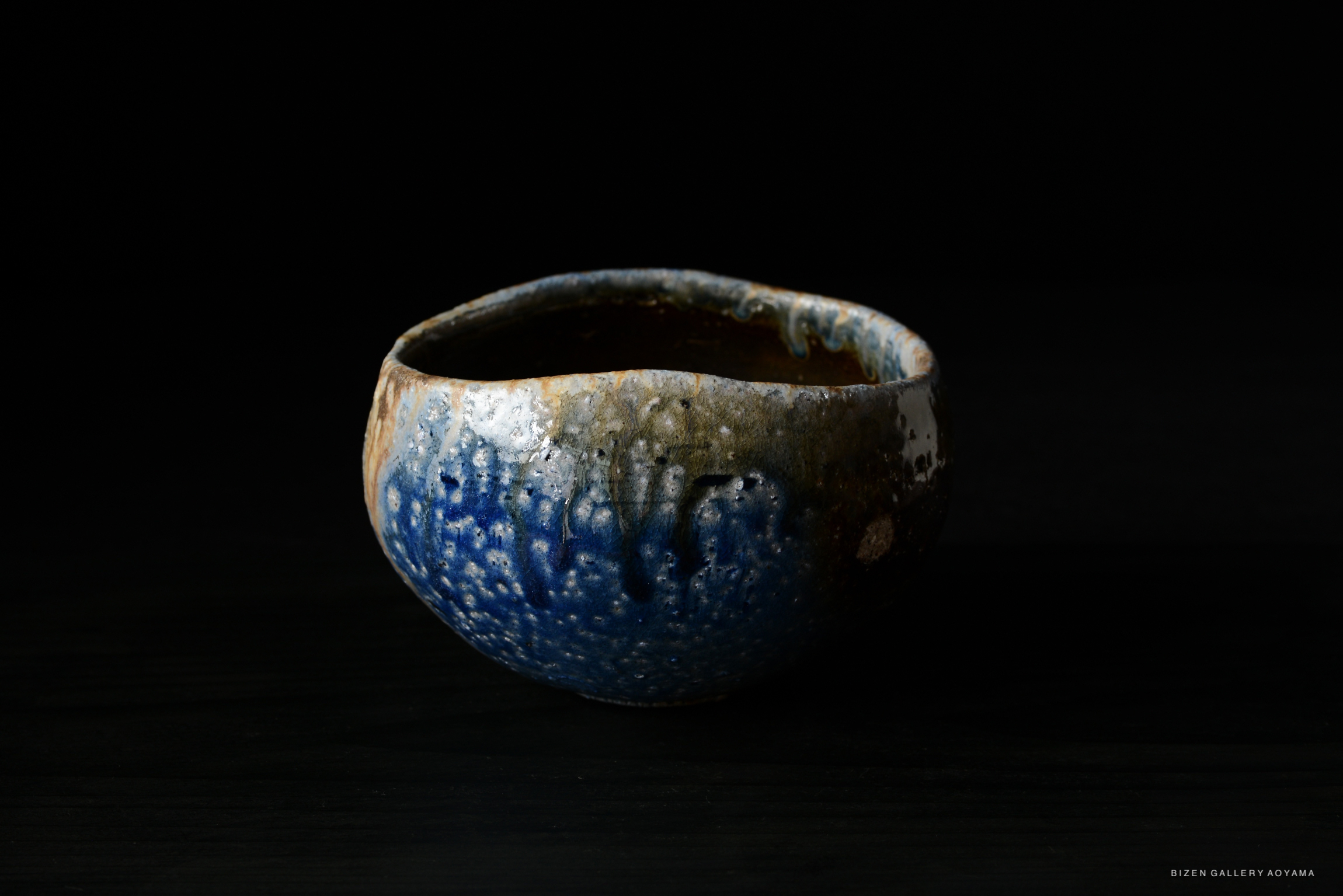 A rustic ceramic bowl with a textured surface, featuring a blend of earthy tones and dark blue accents, set against a black background.