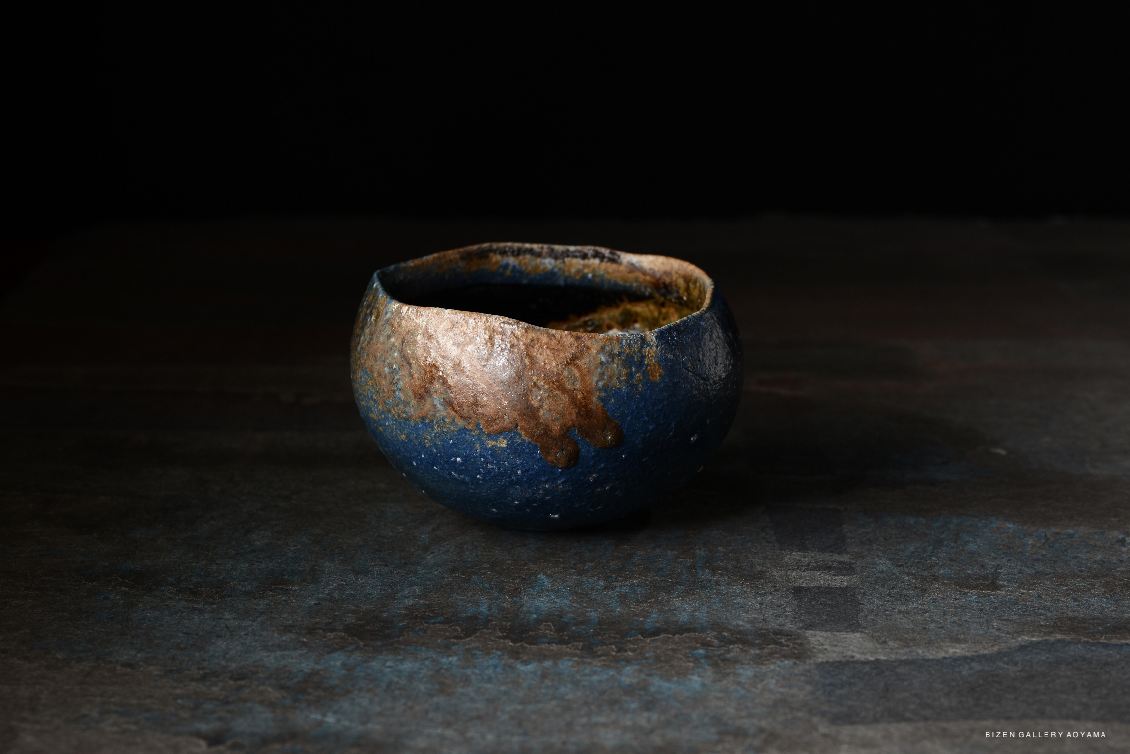 A close-up of a handcrafted ceramic bowl with a textured surface in blue and earthy tones, set against a dark background.