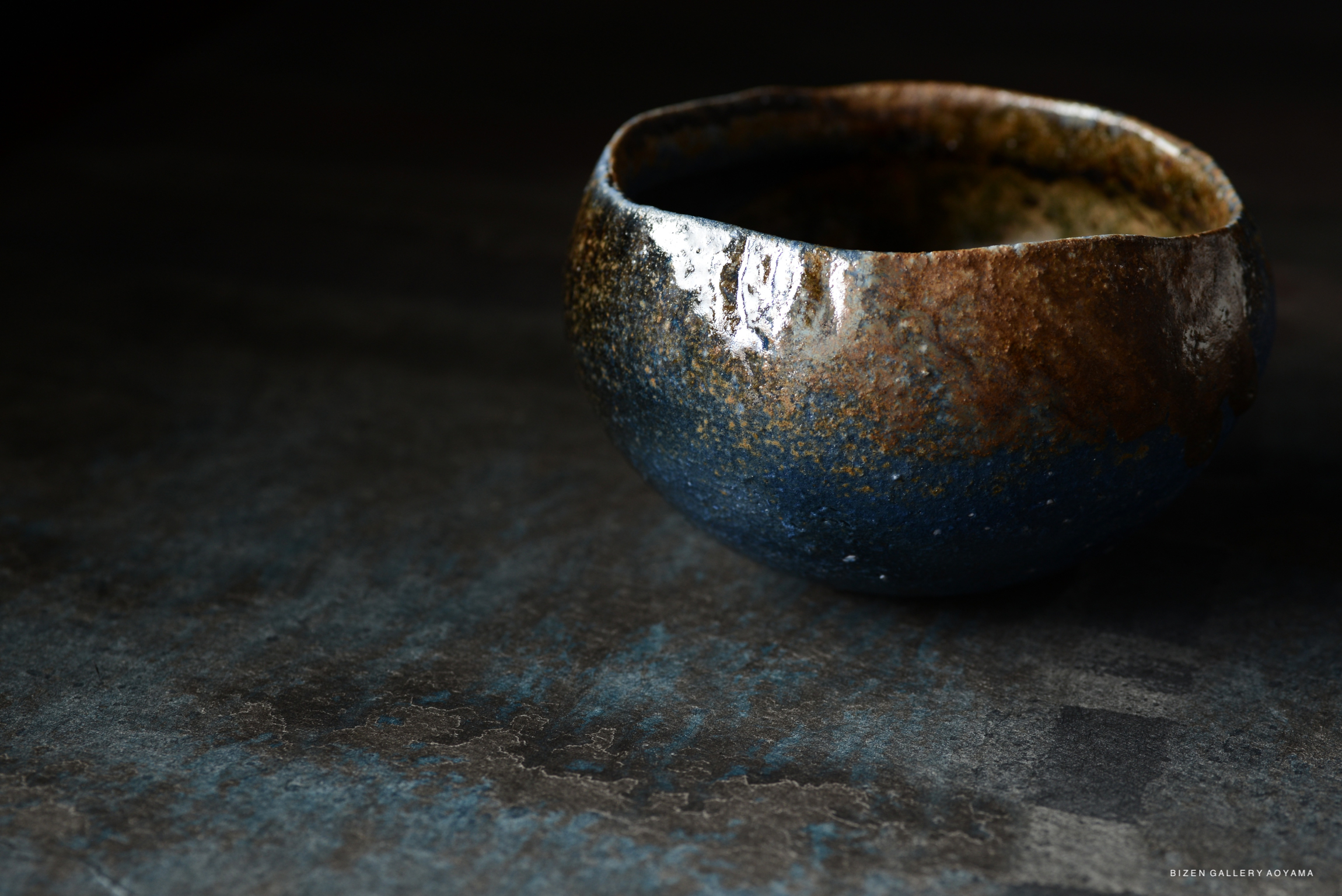A close-up of a handcrafted pottery bowl with a textured surface featuring a blend of dark blue and earthy tones, set against a dark background.