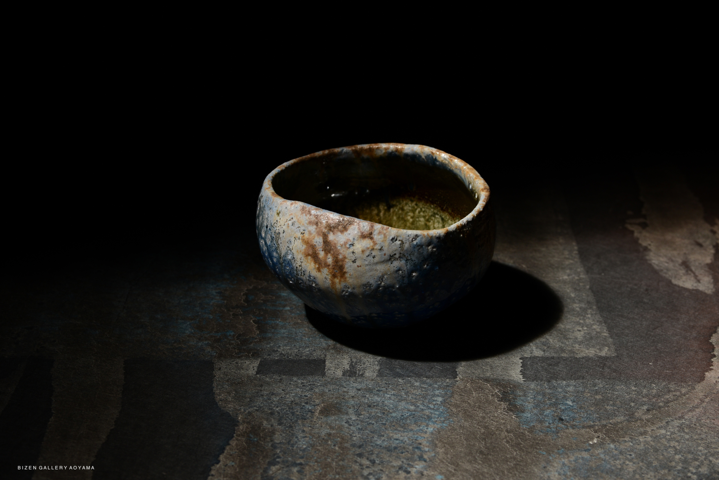 A handcrafted ceramic bowl with a textured surface, exhibiting earthy tones and a glossy finish, placed on a dark surface with subtle lighting.
