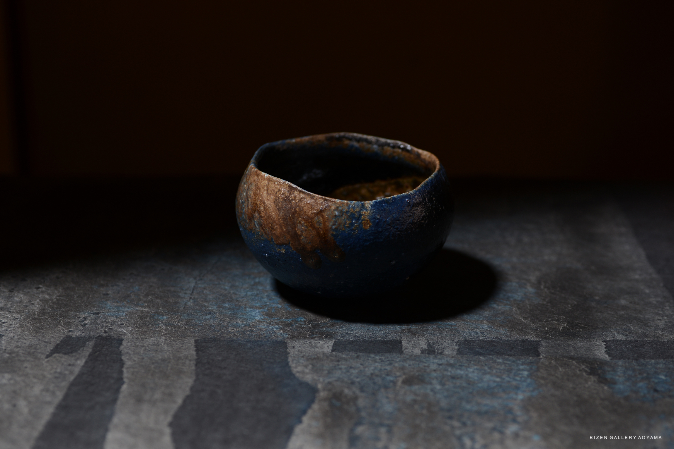 A small, rustic ceramic bowl with a dark blue exterior and a textured surface, placed on a dark gray table. The bowl has a brown rim and is partially illuminated, highlighting its unique glaze.