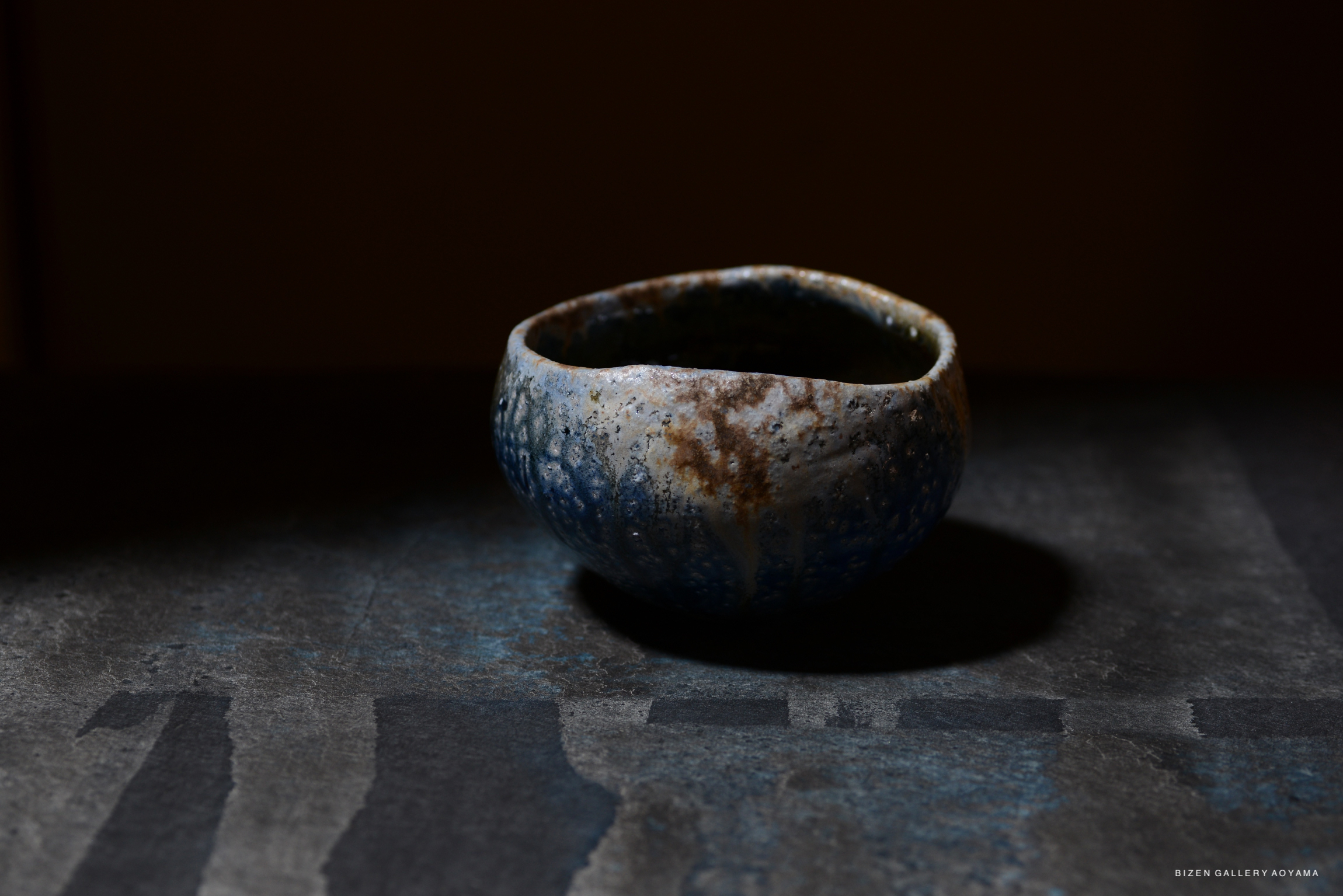 A handcrafted ceramic bowl with a textured surface, featuring shades of blue and specks of brown, placed on a dark table against a blurred background.