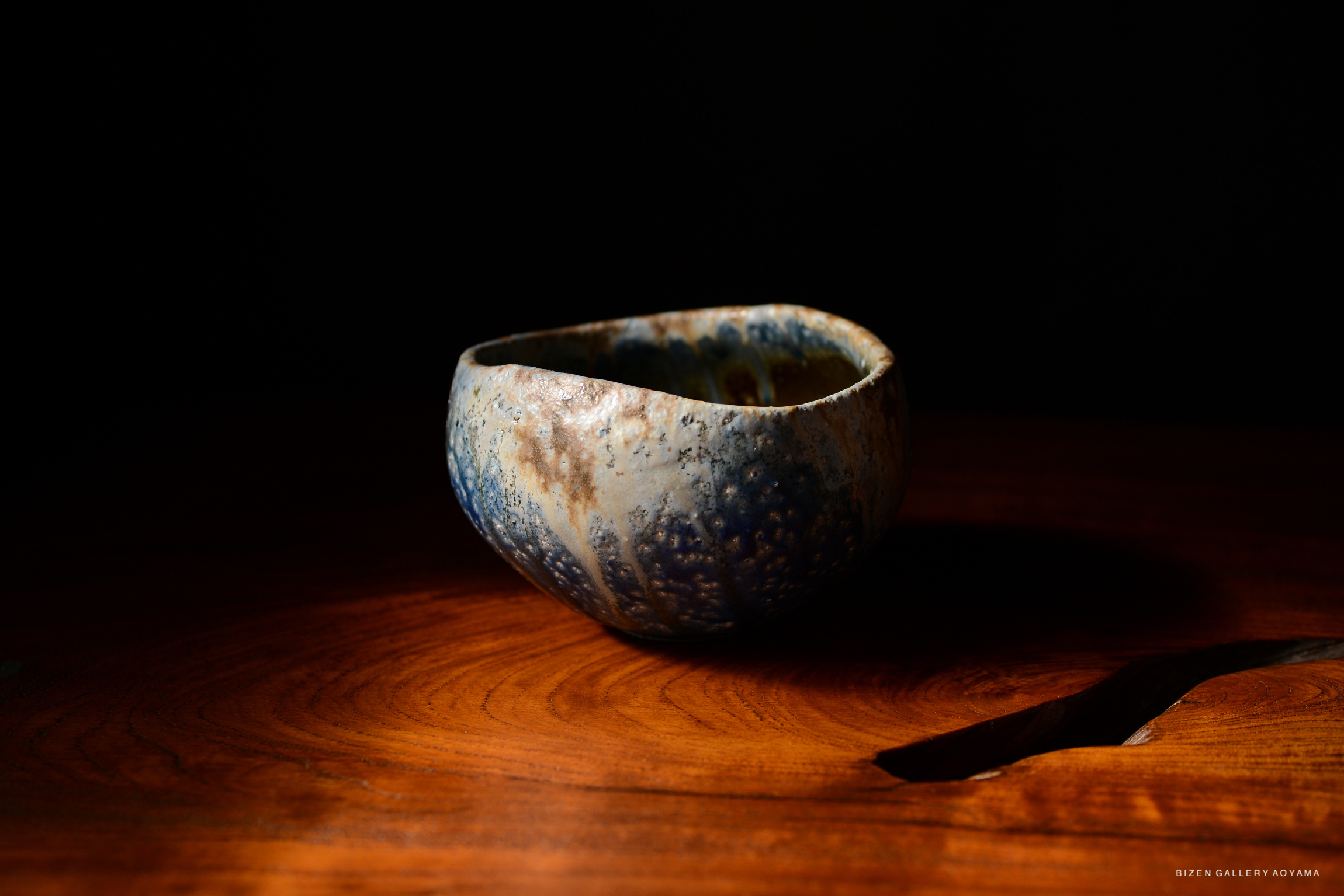 A handcrafted ceramic bowl with a textured surface and blue hues, placed on a wooden table against a dark background.