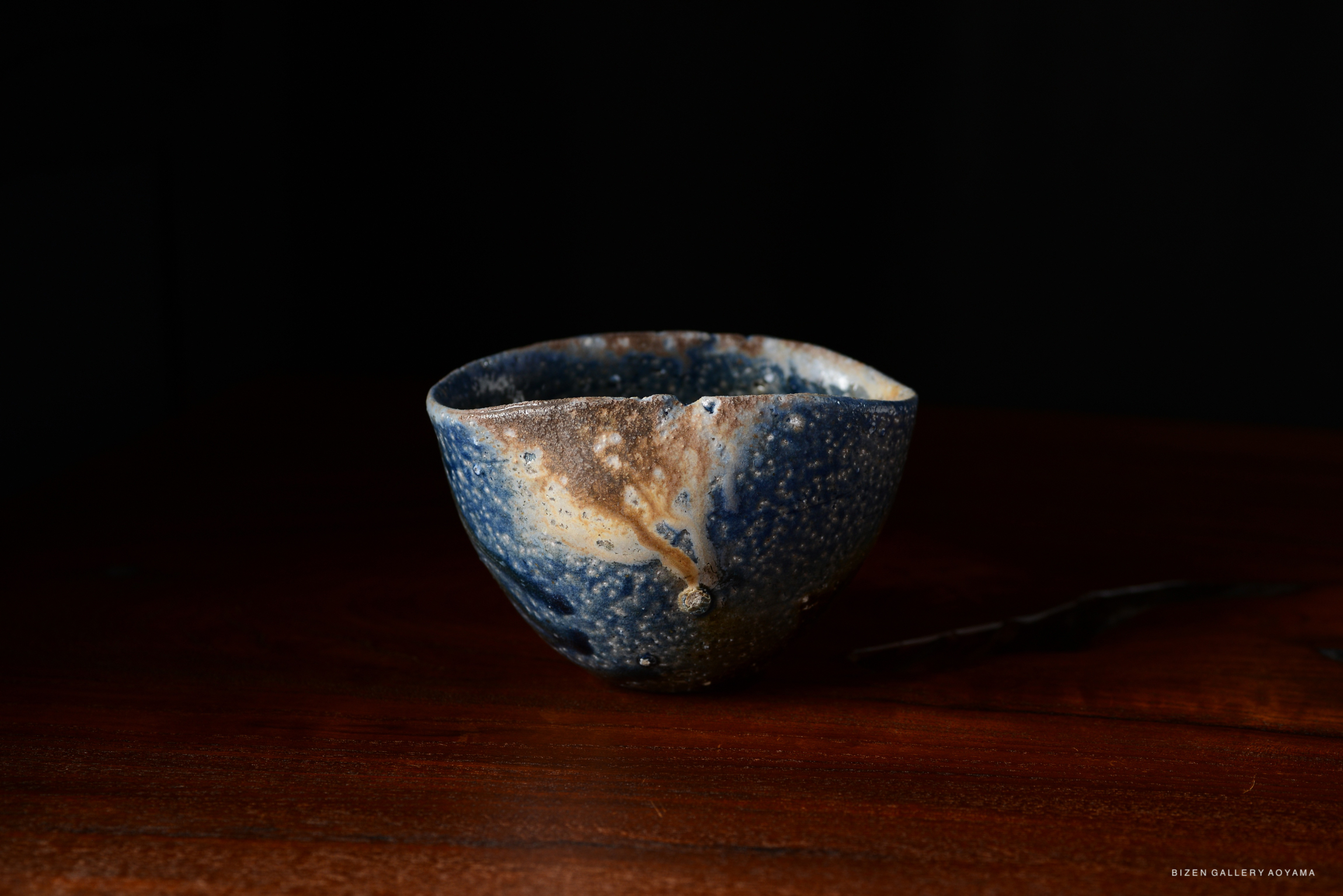 A handcrafted ceramic bowl with a textured surface, featuring various shades of blue and earthy tones, placed on a wooden table against a dark background.