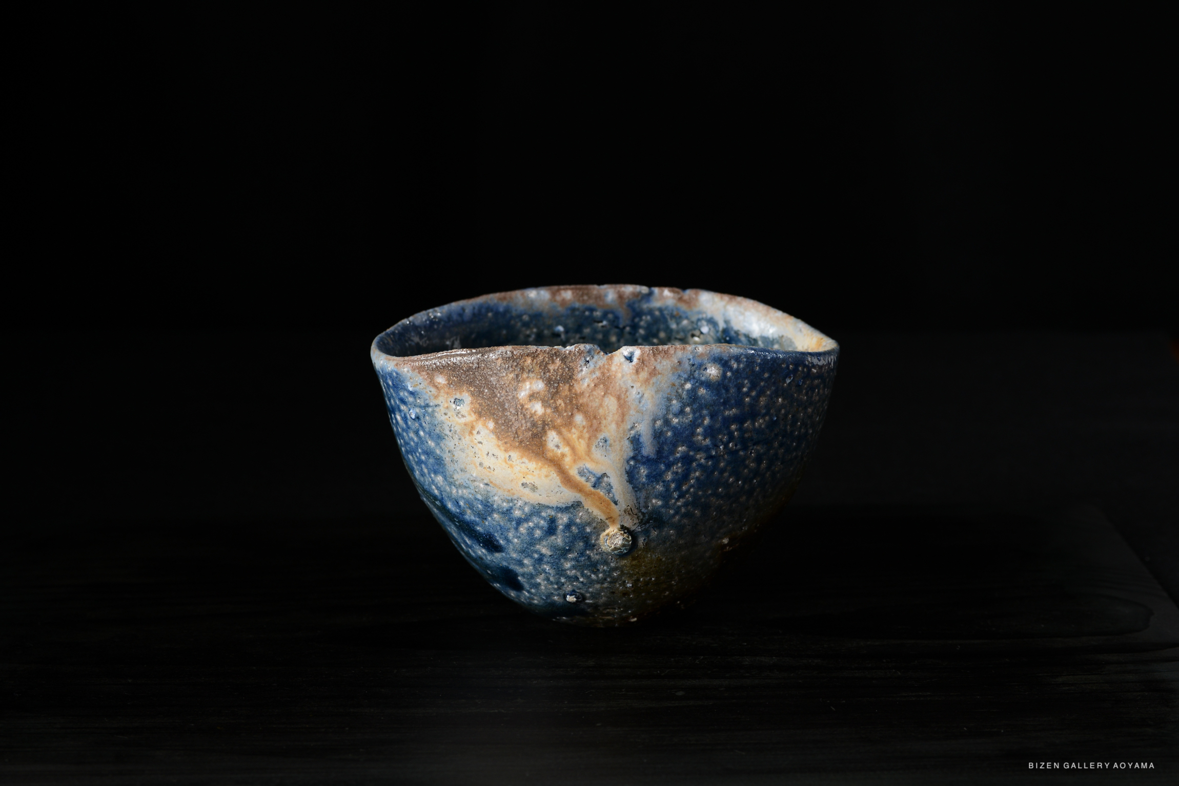 A handcrafted ceramic bowl with a textured blue and brown glaze, set against a black background.