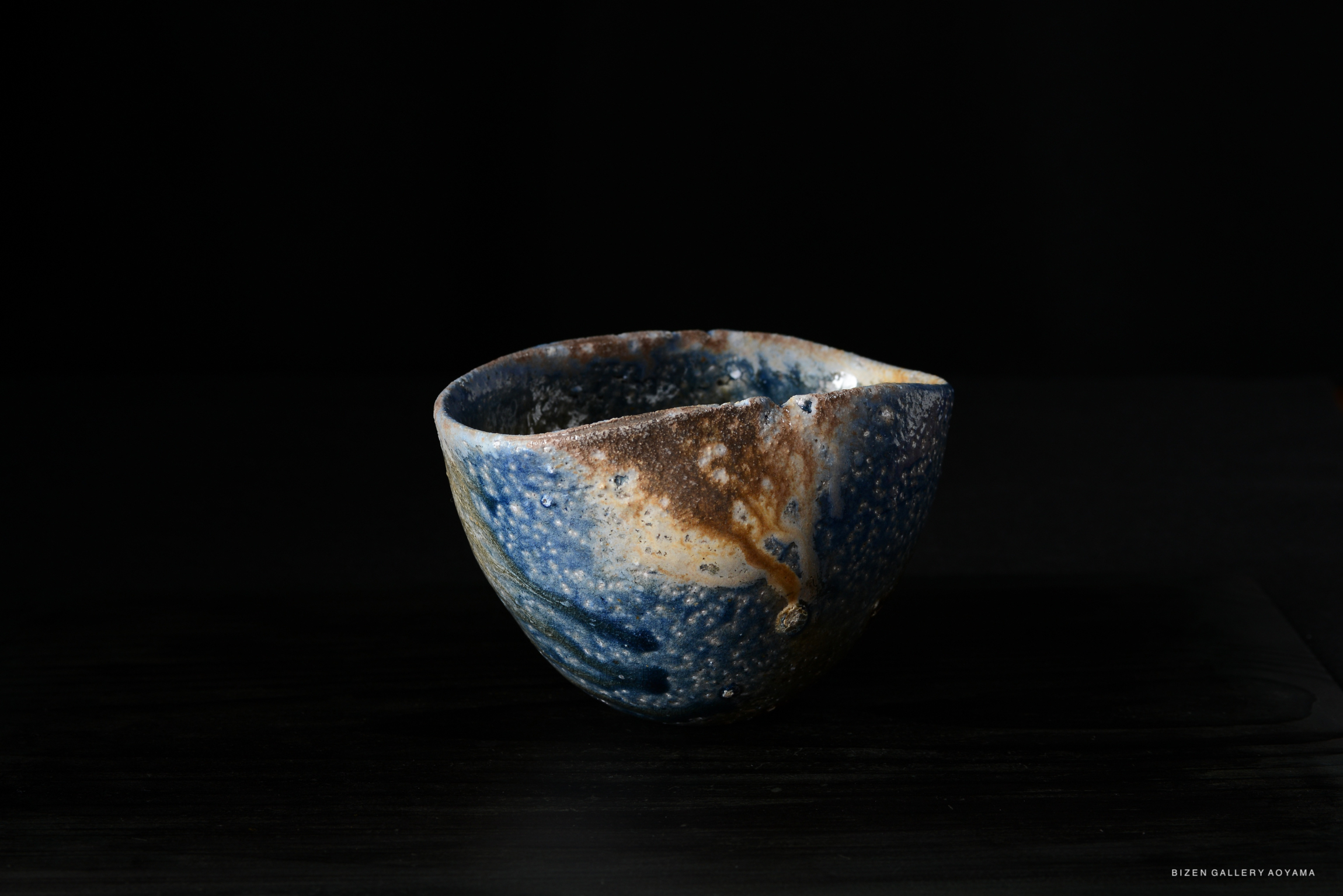 A close-up of a uniquely shaped ceramic bowl with a textured surface, featuring shades of blue, brown, and cream against a dark background.