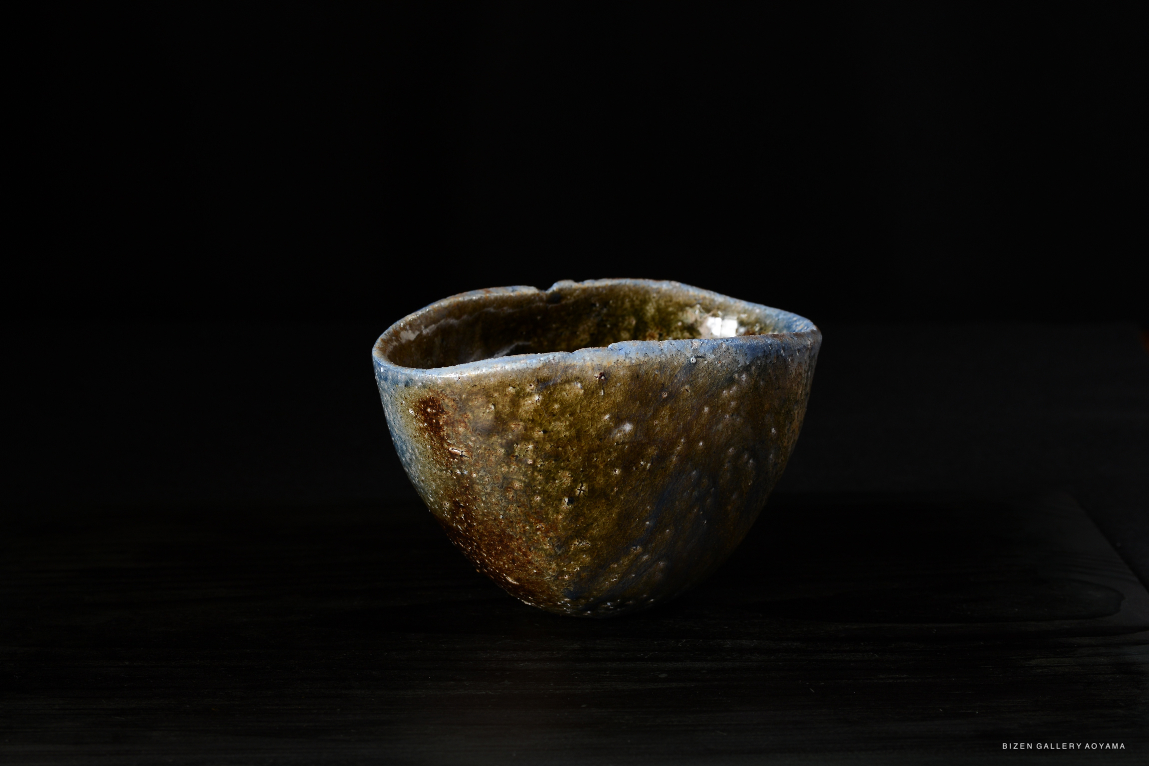 A unique, handcrafted ceramic bowl with a textured, earthy glaze in shades of brown and blue, set against a dark background.