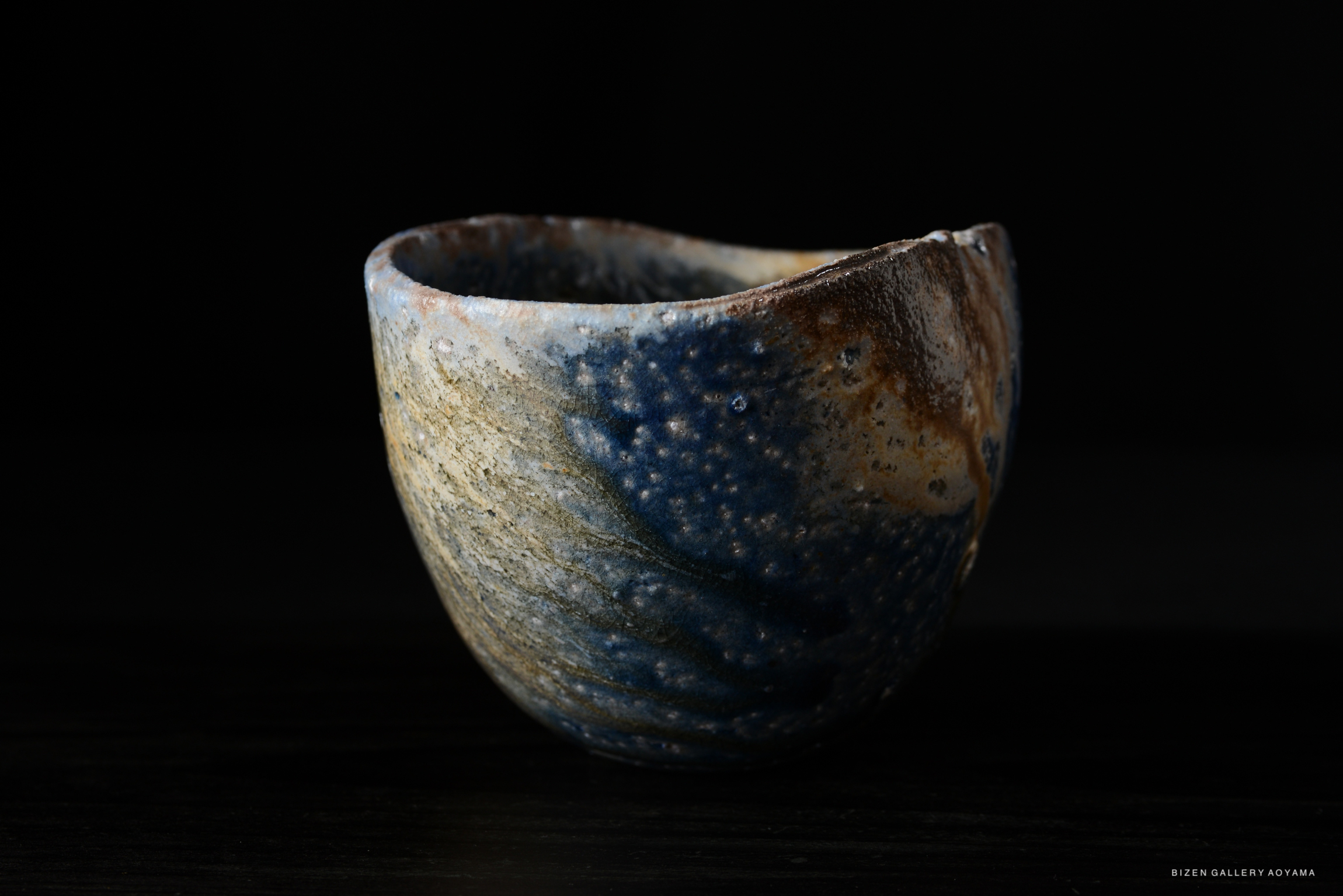 A handcrafted ceramic cup with a textured surface featuring blue and brown glazes, set against a dark background.