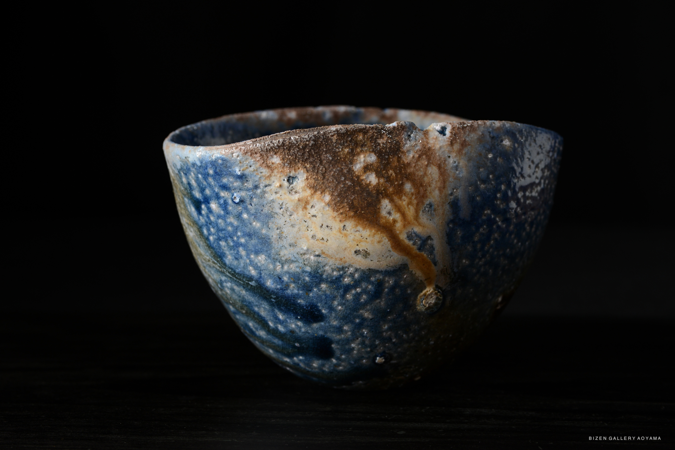 A close-up image of a uniquely shaped ceramic bowl with a blue and brown glaze, set against a black background.