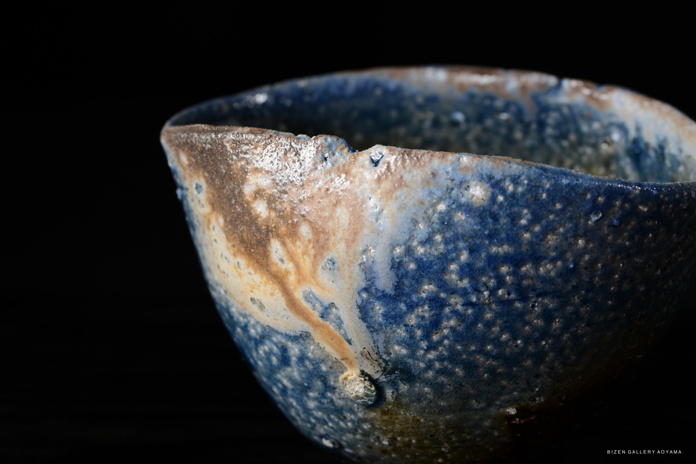 Close-up of a handcrafted ceramic bowl featuring a textured surface with blue, brown, and beige hues against a dark background.