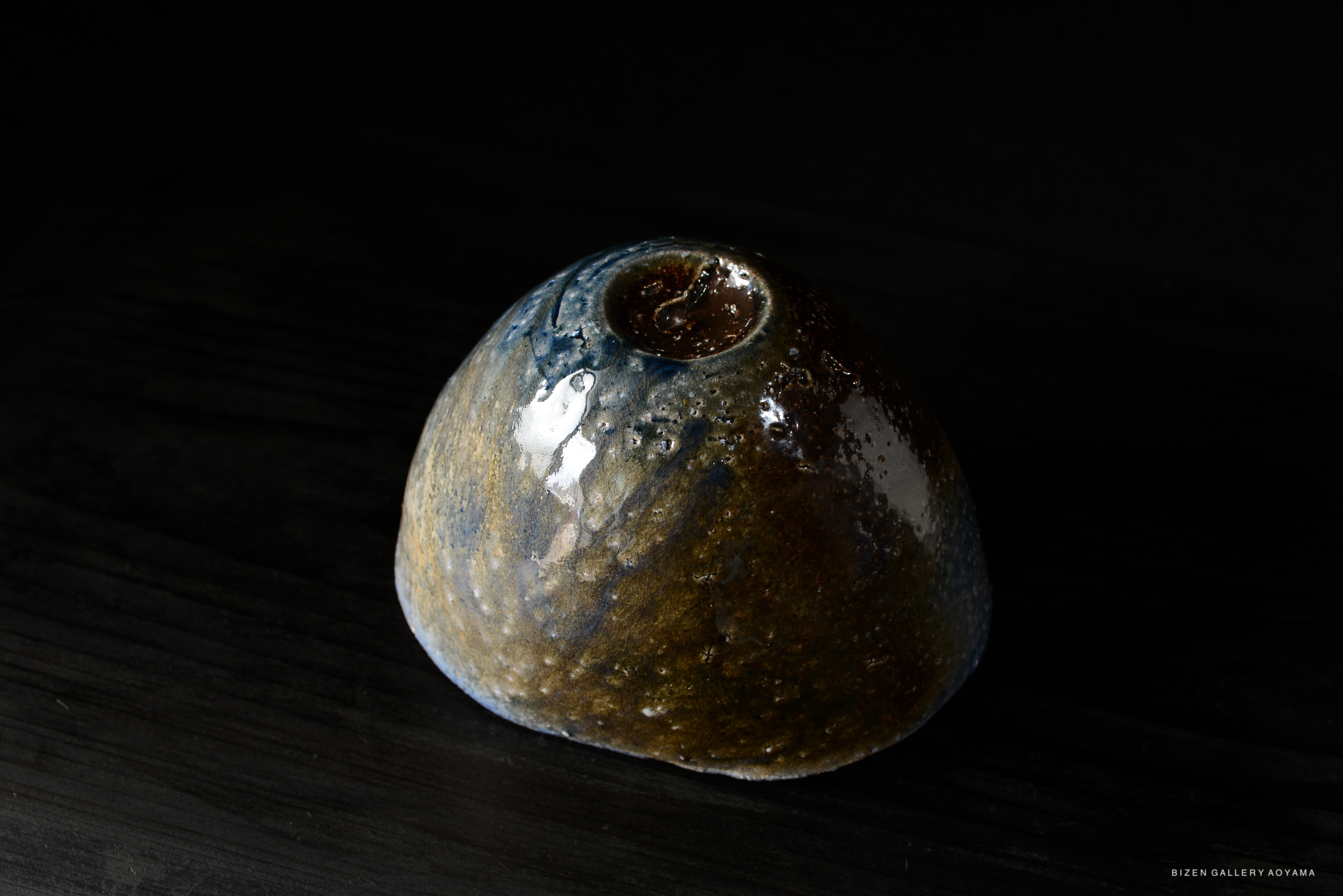 A handcrafted ceramic vase with a unique, textured surface and a blend of earthy colors, resting on a dark wooden surface.