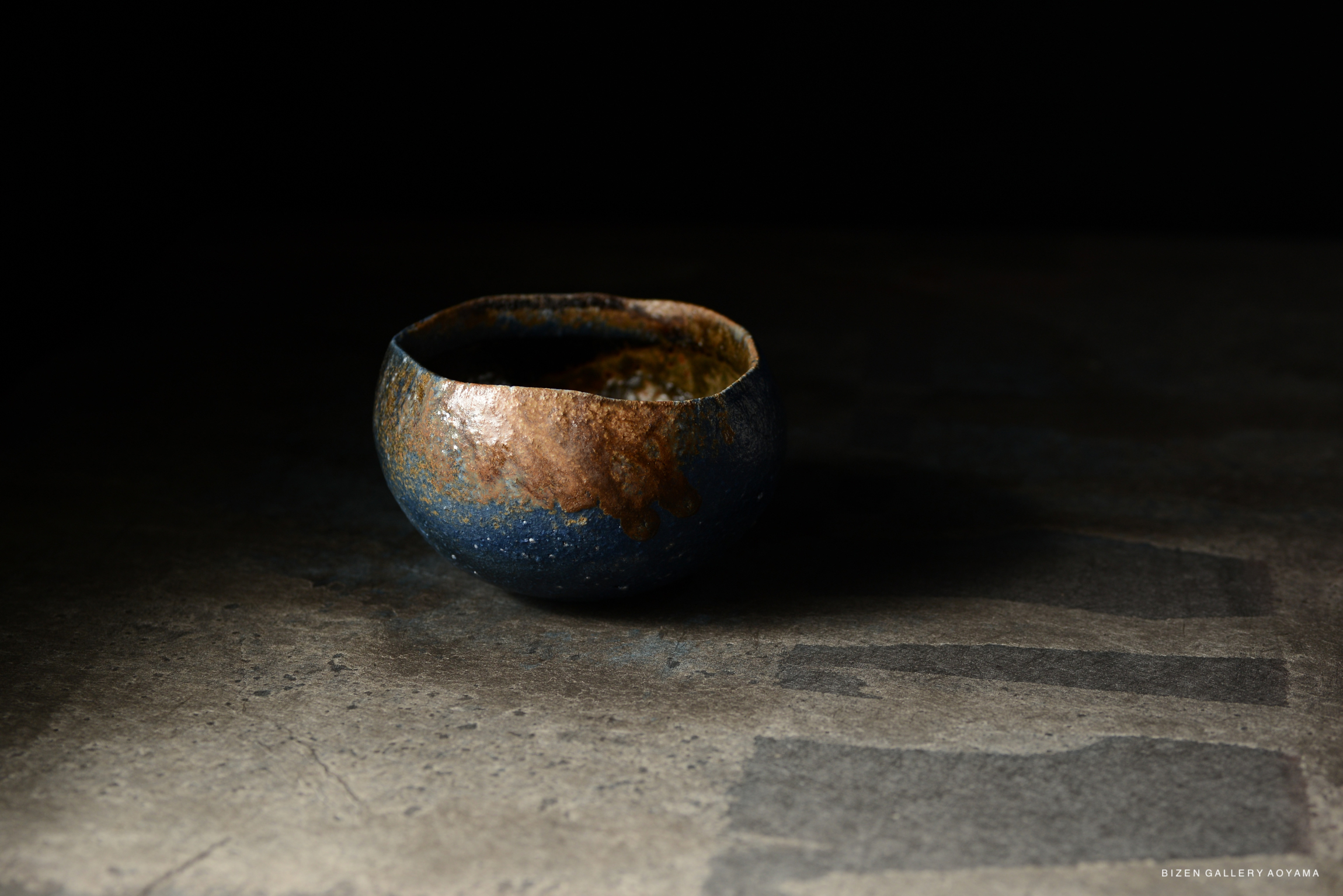A handcrafted ceramic bowl with a dark blue and earthy brown glaze, showcasing organic textures and colors, resting on a textured surface.