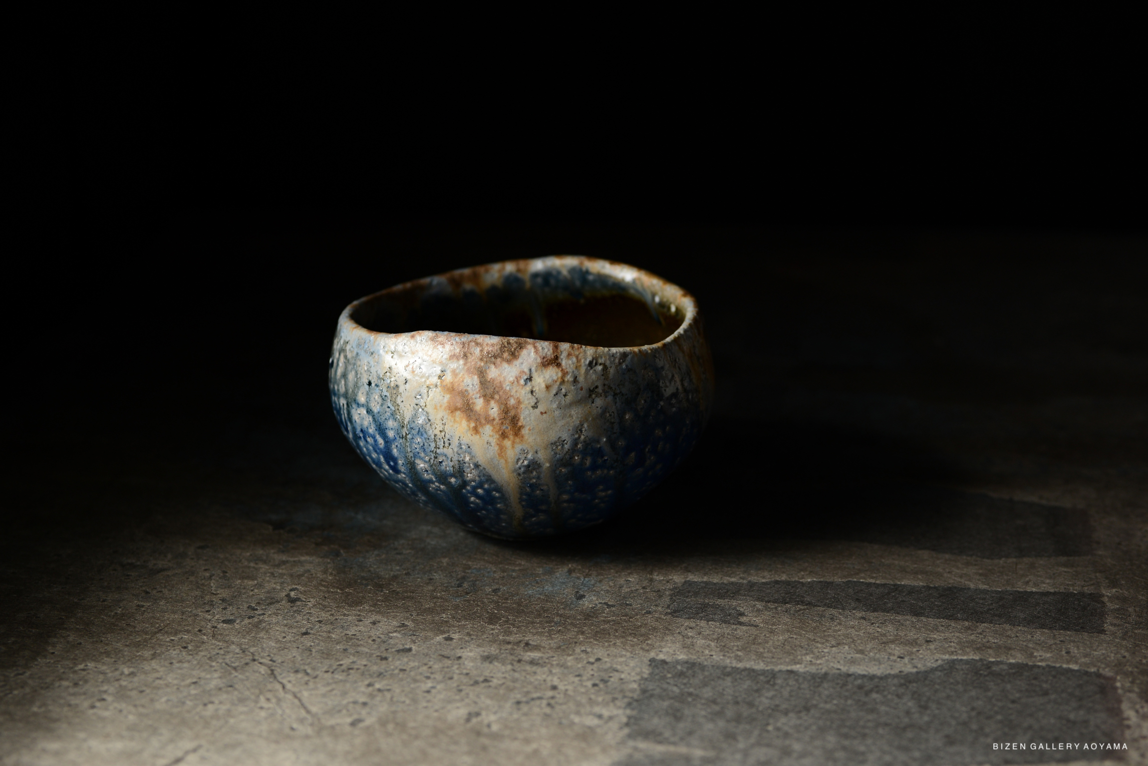 A handcrafted ceramic bowl with a textured blue and earthy color glaze, sitting on a dark surface.