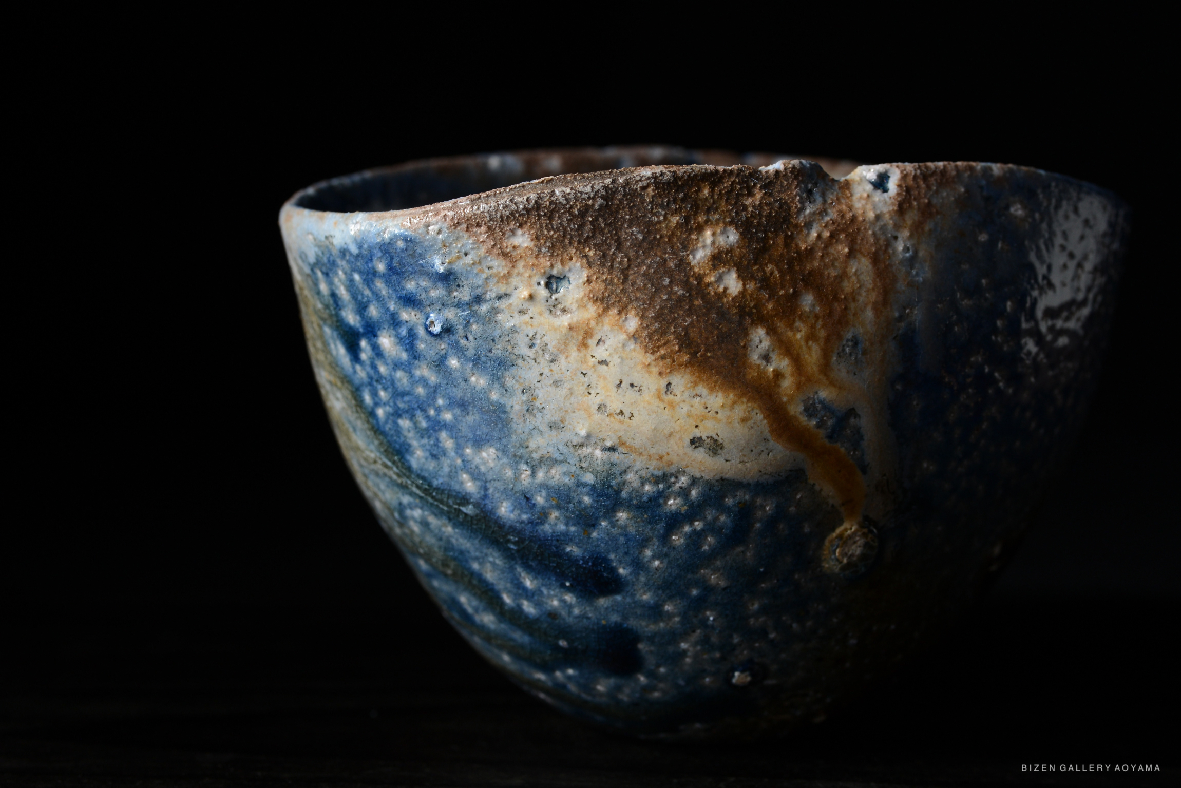 A close-up image of a beautifully glazed ceramic bowl featuring shades of blue and brown, with a textured surface, set against a dark background.