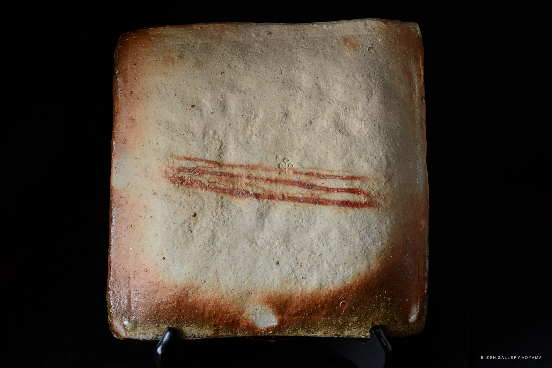 A flat, rectangular Bizen pottery piece with a textured surface featuring reddish-brown markings, displayed on a stand against a black background.