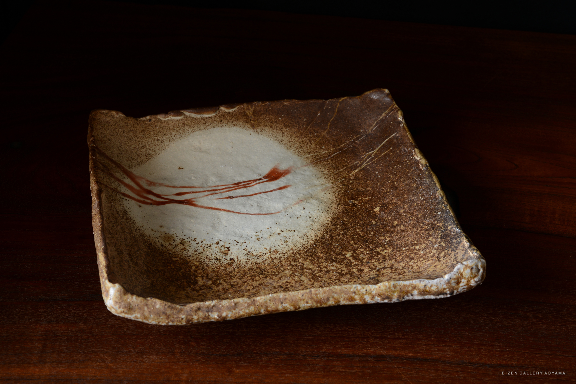 A handcrafted Bizen pottery plate with a textured, irregular shape and subtle red brushstroke decorations on a pale center.