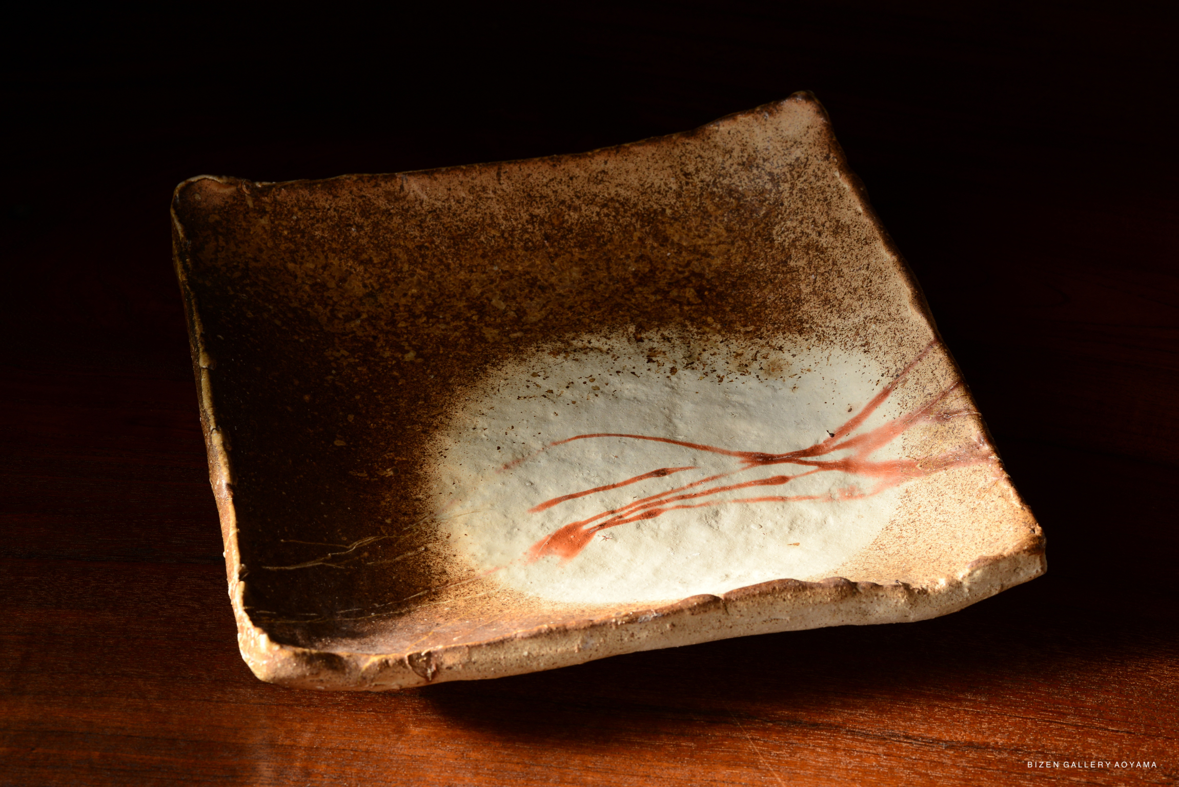 A square Bizen pottery plate with earthy tones and a white center marked by red brushstrokes, set against a dark background.
