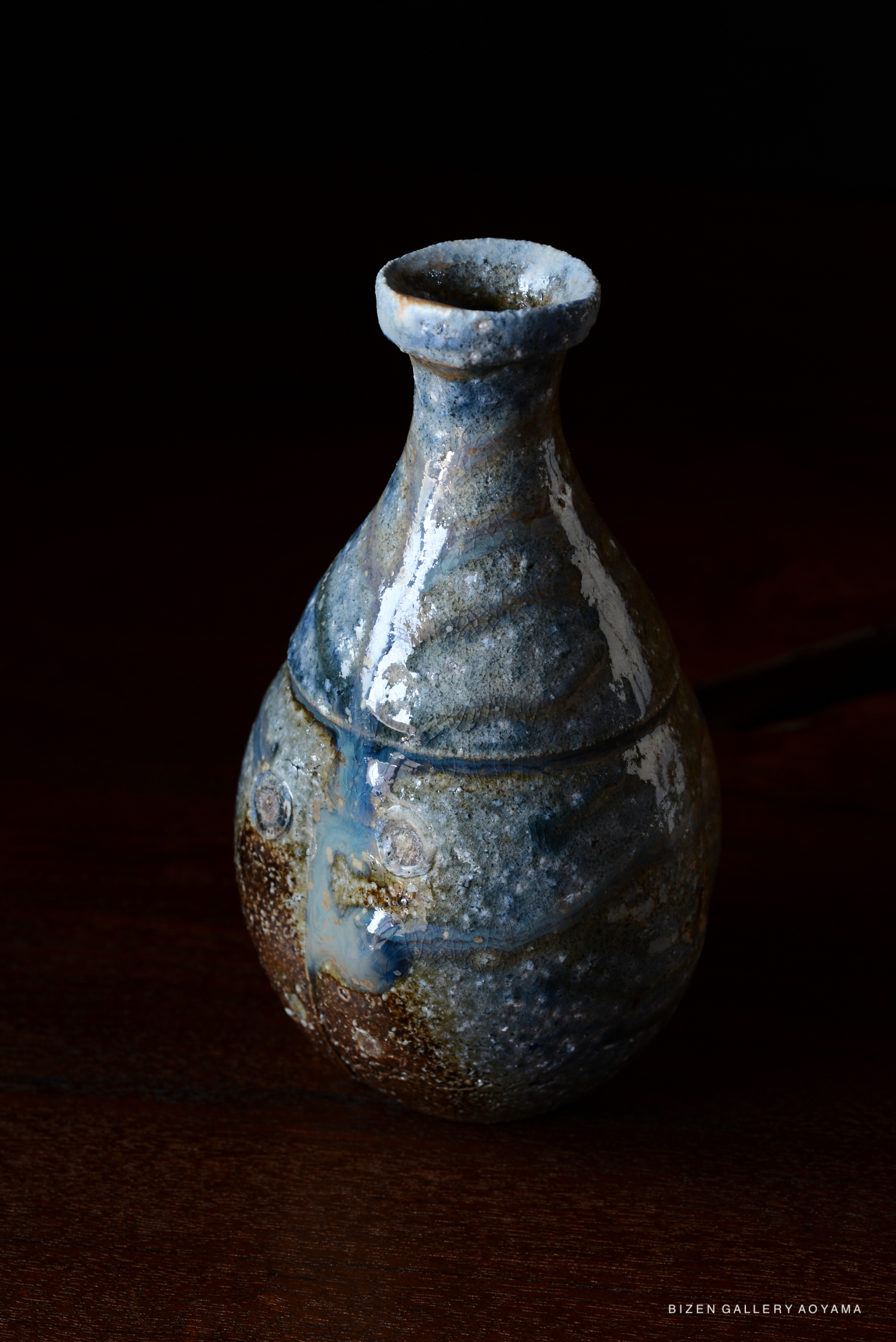 A blue-toned ceramic sake bottle with a rounded body and narrow neck, displayed on a wooden surface with a dark background.