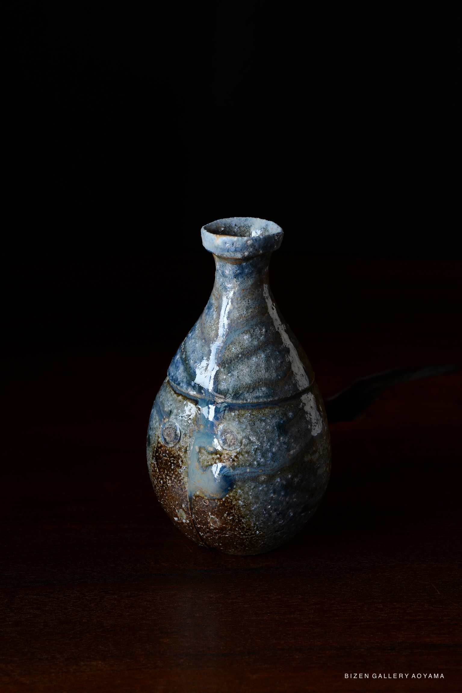A handcrafted Bizen pottery vase with a smooth, glossy finish featuring shades of blue and brown, set against a dark background.