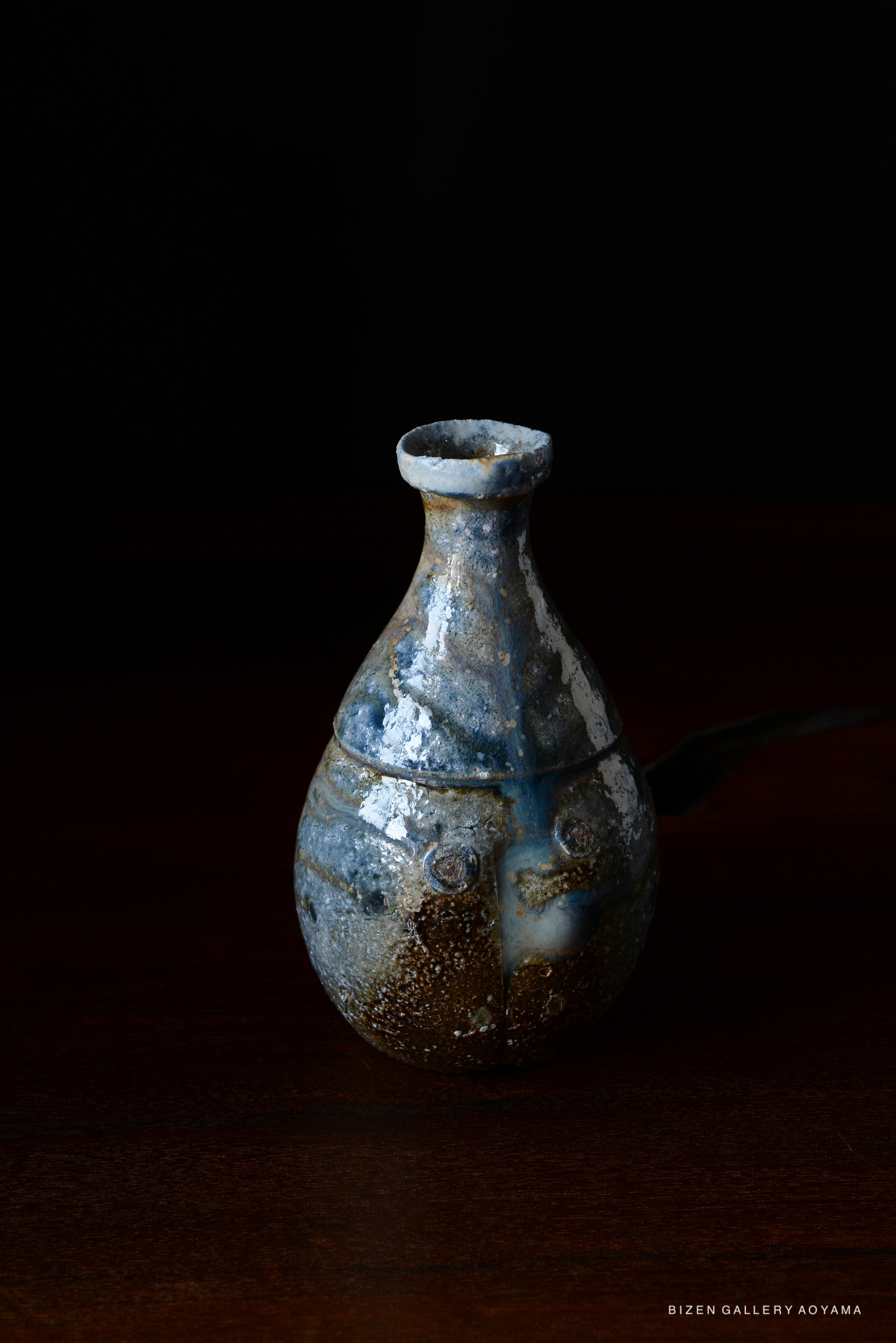 A ceramic Bizen pottery sake vessel with a unique blue and brown glaze, featuring a rounded shape and a narrow neck, set against a dark background.