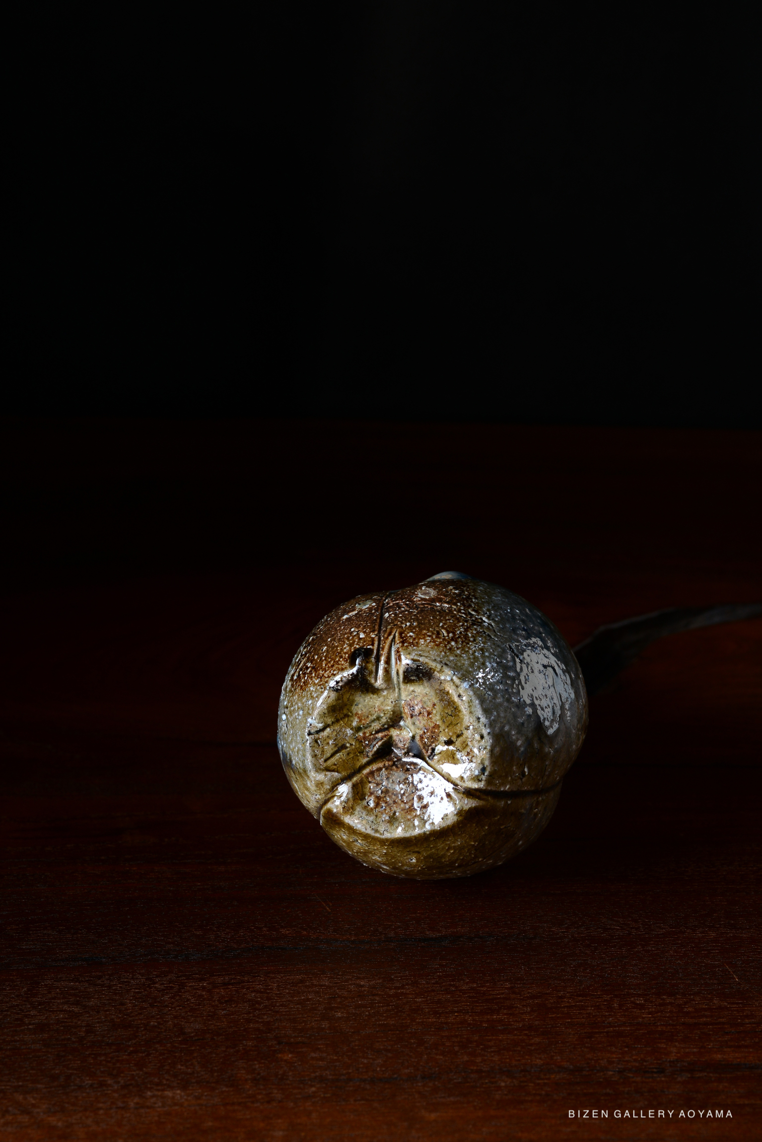 A close-up of a unique Bizen pottery piece, resembling a round, textured object with a metallic sheen, set against a dark background.