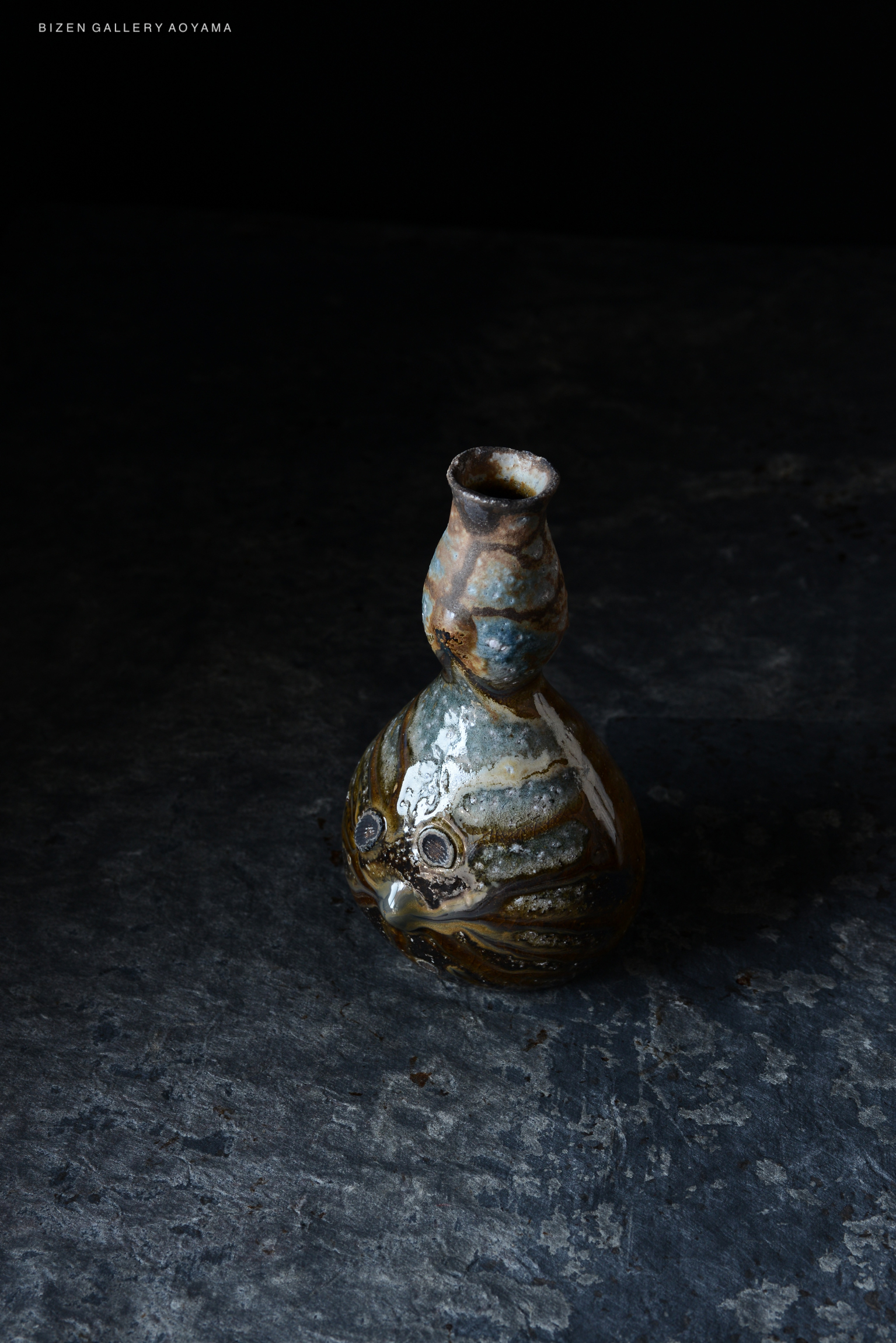 A unique ceramic vase with a curved shape, featuring a textured surface and a mix of earthy colors, placed against a dark backdrop.