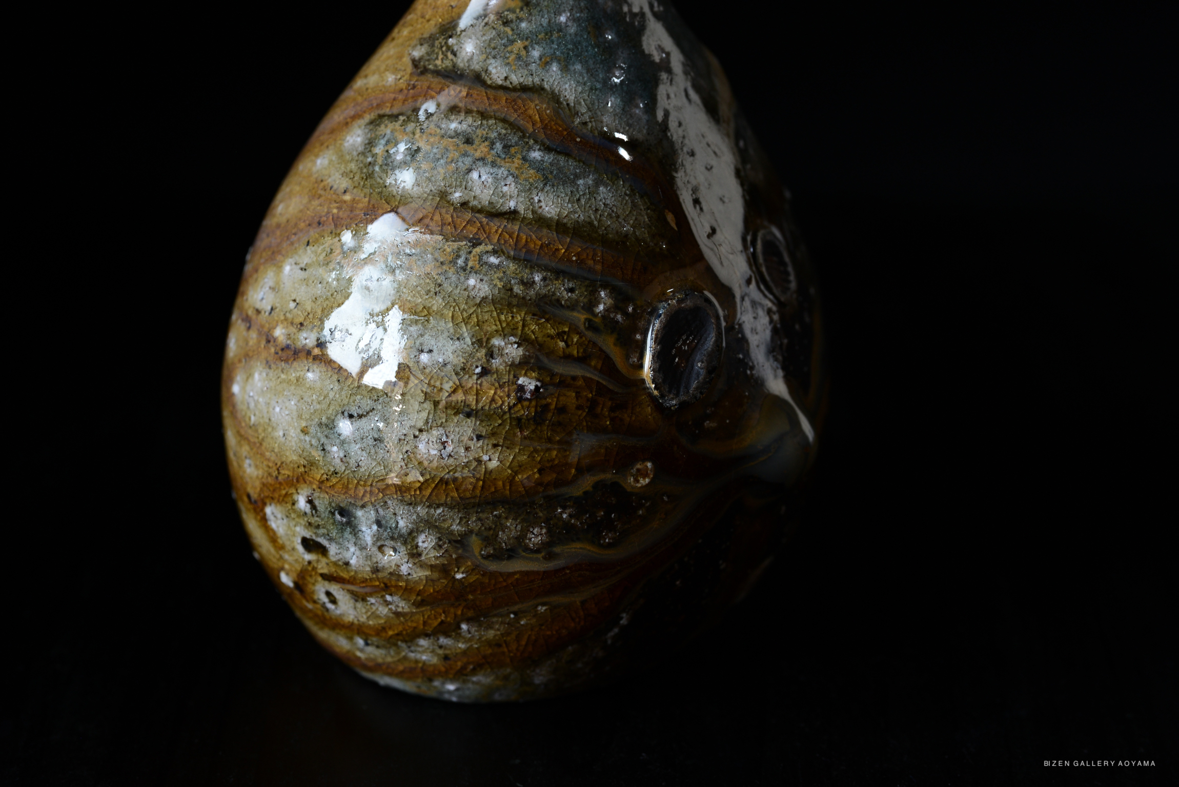 A close-up of a uniquely shaped ceramic object with a glossy finish and intricate textures, placed against a dark background.