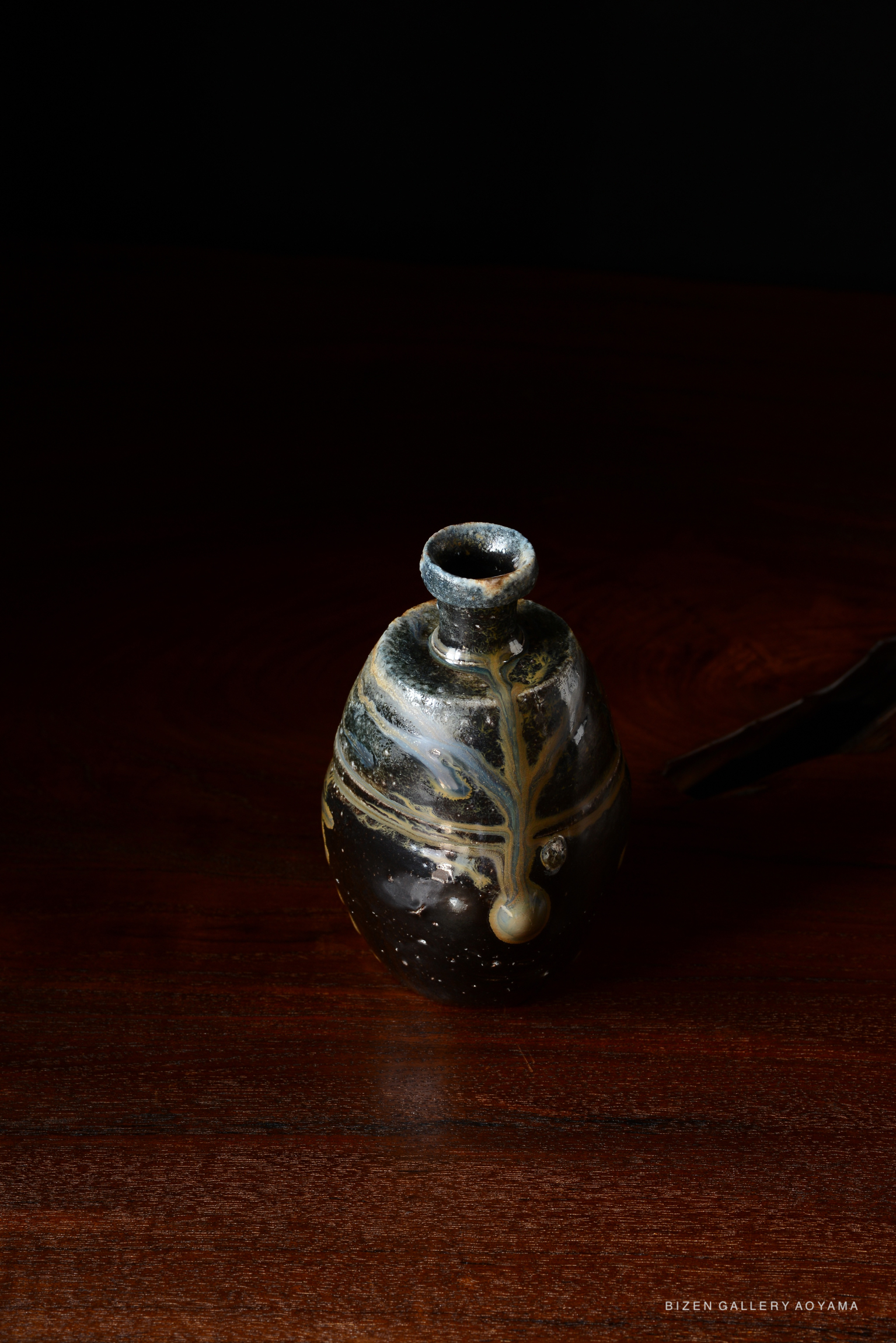A handmade black ceramic Tokkuri (sake flask) with intricate glaze patterns, displayed on a dark wooden surface.