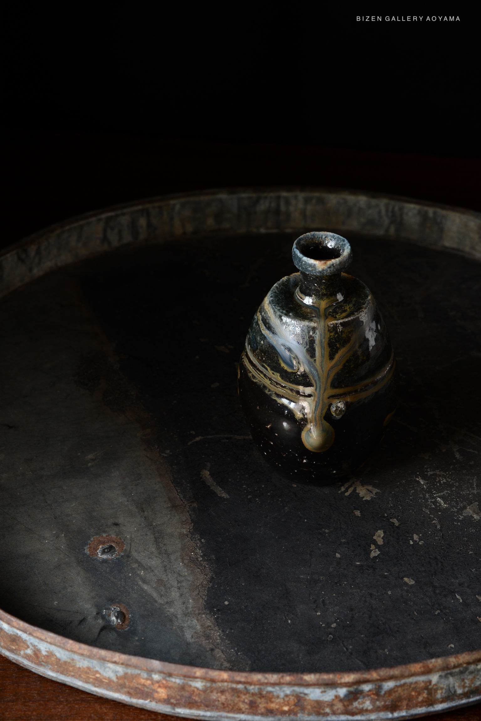 A close-up image of a traditional Bizen pottery Tokkuri placed on a textured, round metal tray against a dark background.